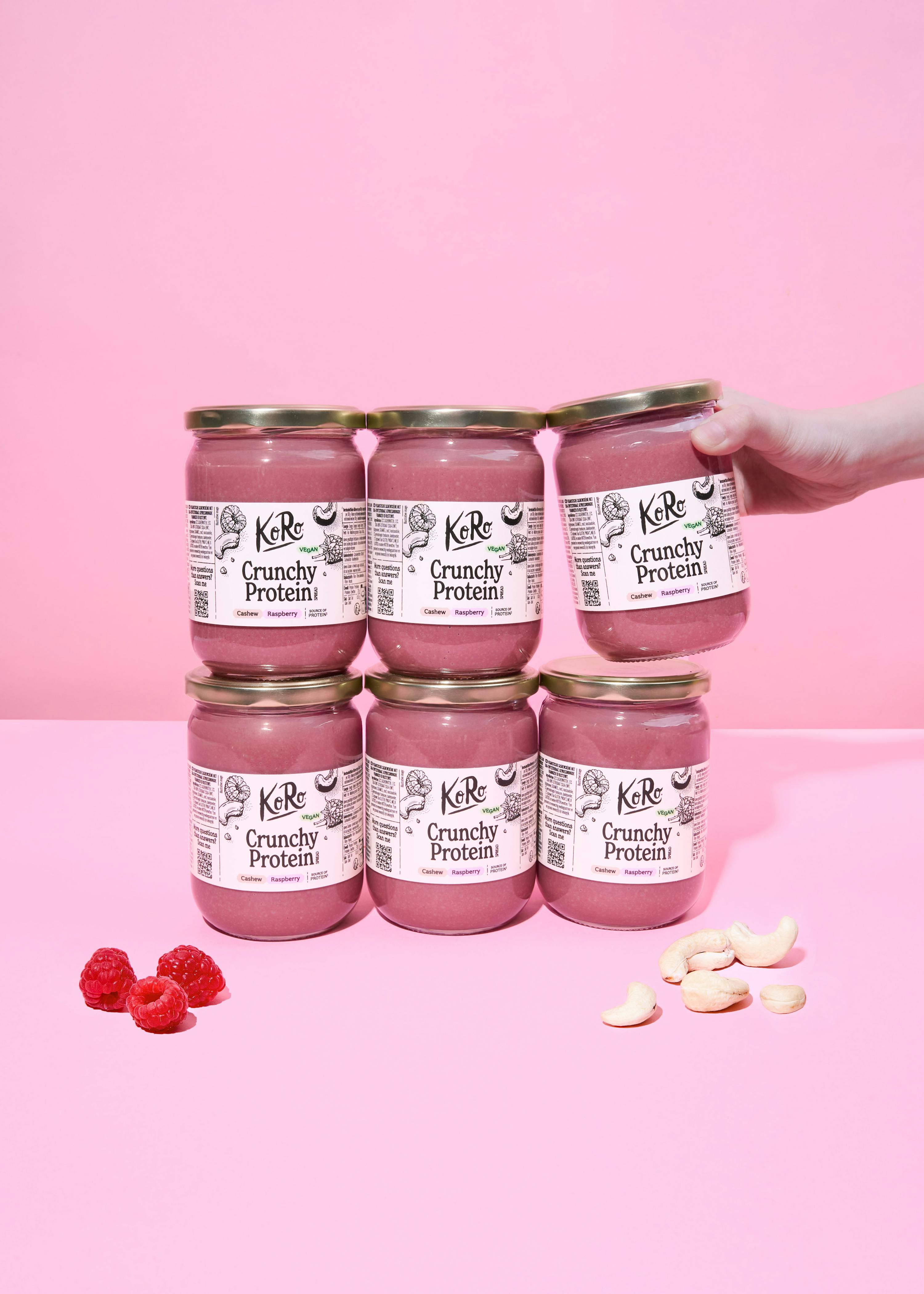 sechs gläser koro crunchy protein cashew-himbeer-aufstrich auf einem pinken hintergrund, wobei ein glas von einer hand gehalten wird und frische himbeeren sowie cashewnüsse daneben liegen.