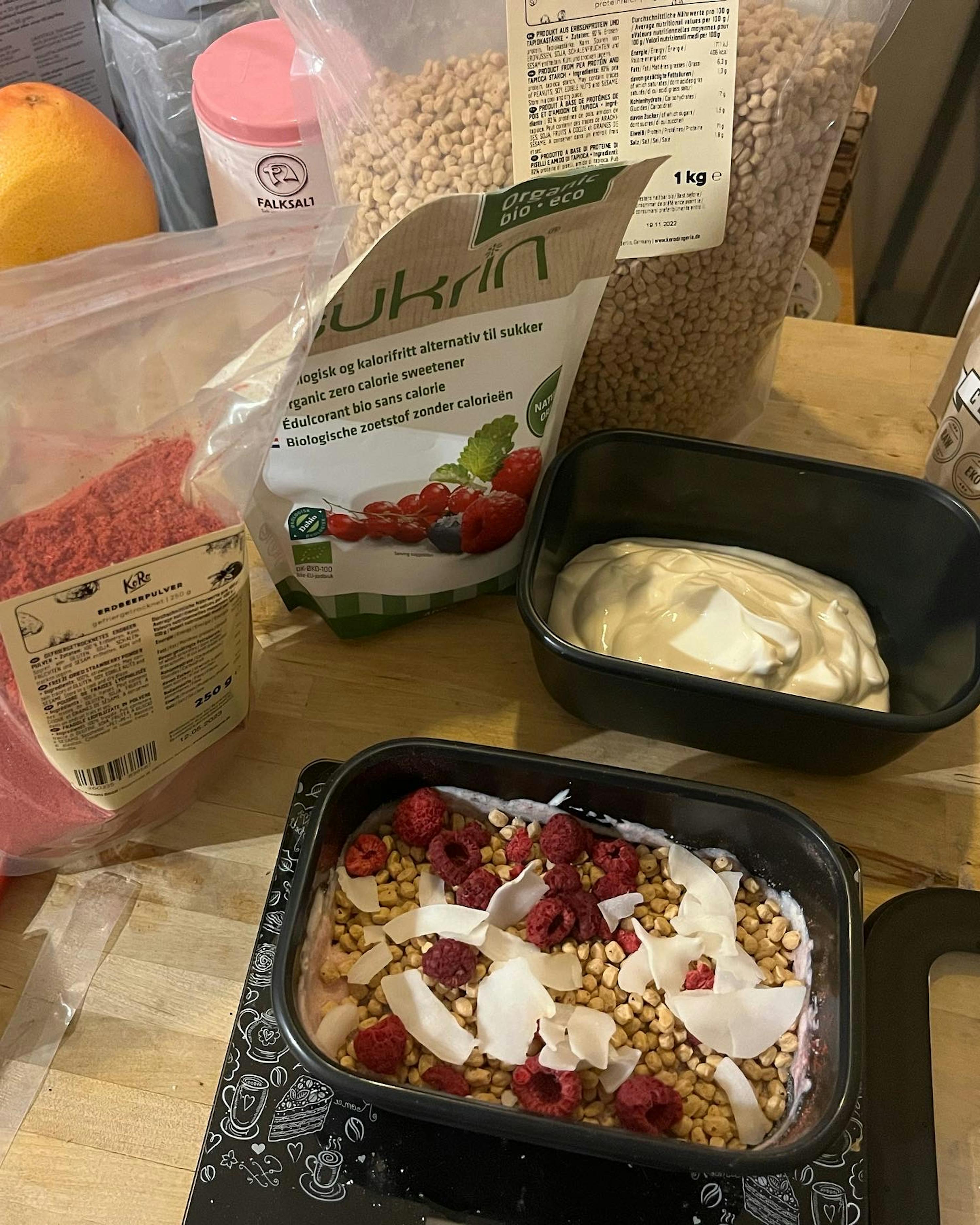 auf einem tisch steht ein container granola mit himbeeren und kokosnuss