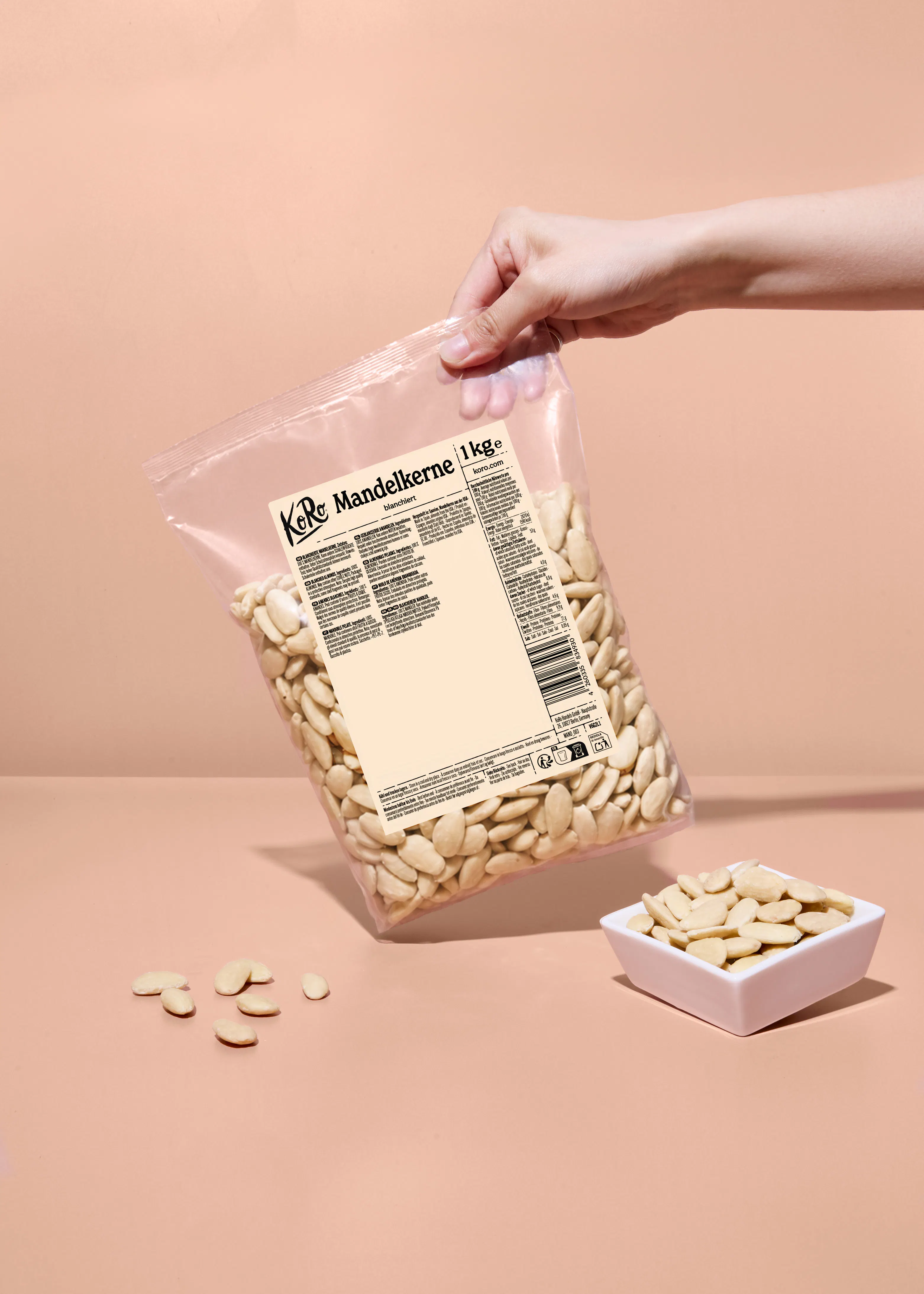 eine hand hält eine transparente tüte mit 1 kg blanchierten mandelkernen der marke koro, während einige mandeln auf einer pfirsichfarbenen oberfläche verstreut sind und eine kleine weiße schale ebenfalls mandeln enthält.