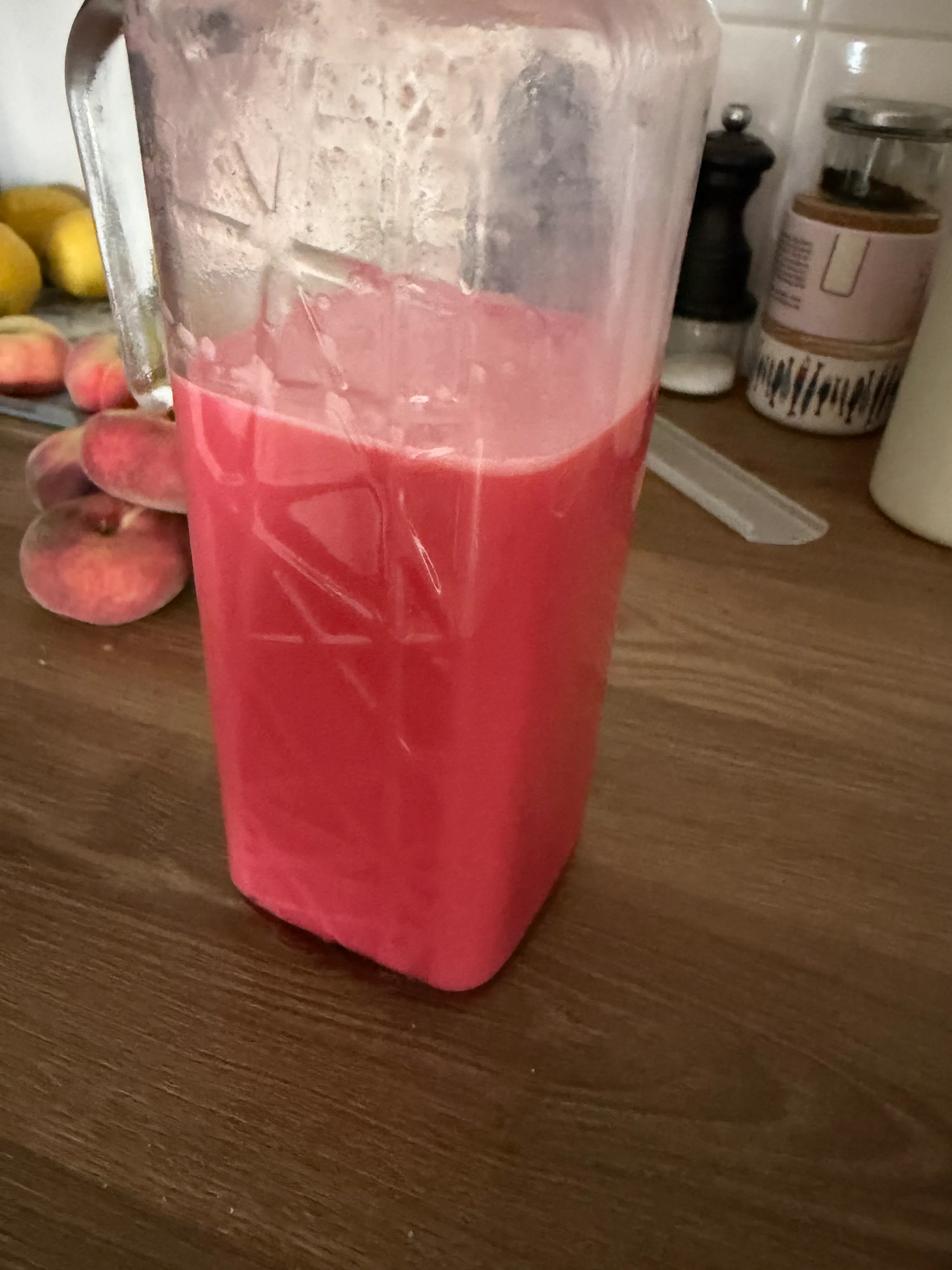 un pichet de mixeur transparent rempli d'un liquide rose, avec des pêches et des citrons sur une surface en bois.