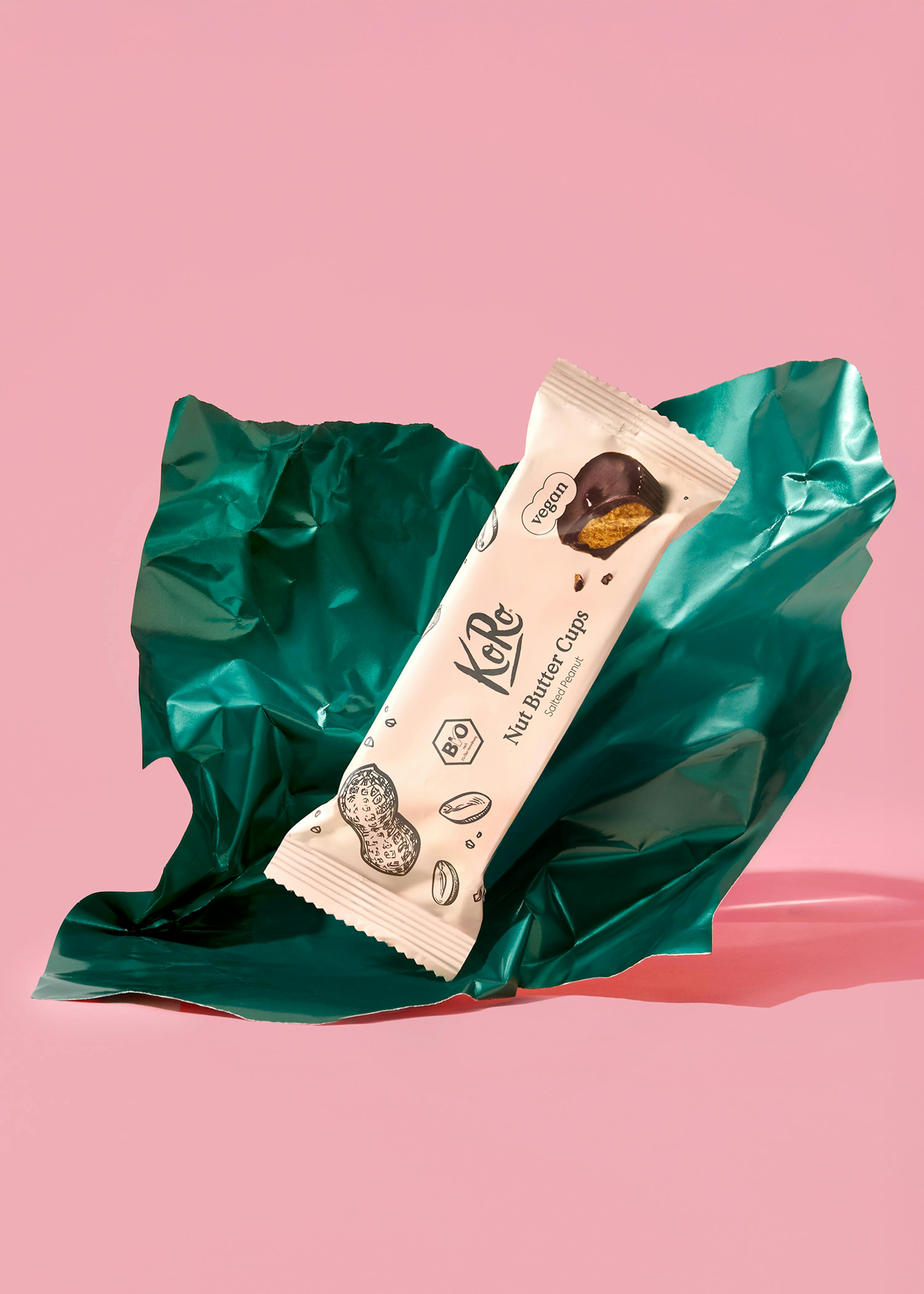 eine packung koro nut butter cups salted peanut, die vegan und bio ist und einen angebissenen erdnussbutter-cup zeigt, liegt auf einer zerknitterten dunkelgrünen verpackung vor einem pinken hintergrund.