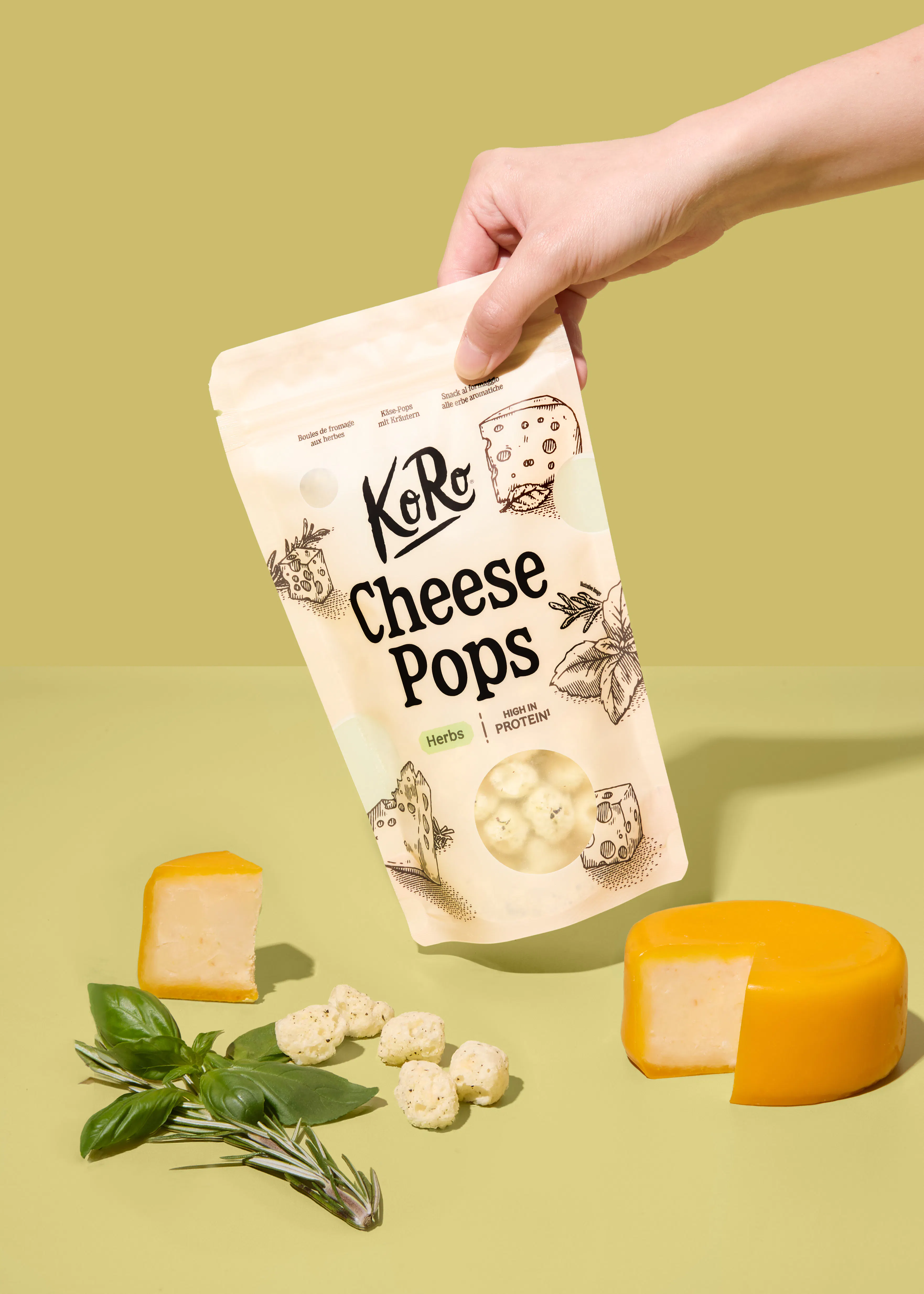 a hand holds a bag of koro herb cheese pops above a surface with fresh basil, rosemary, and two wedges of yellow cheese.