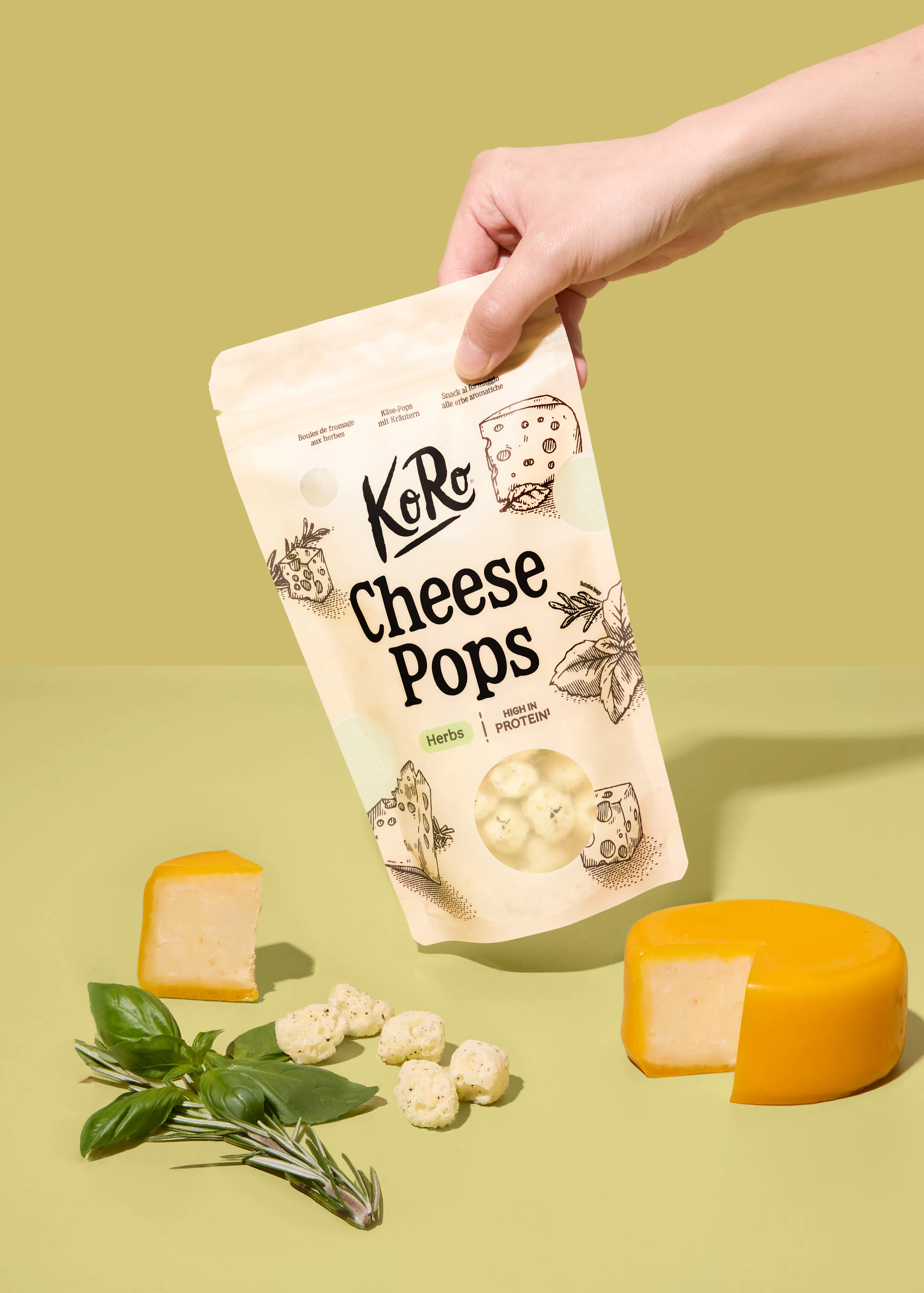 una mano tiene una confezione di snack koro cheese pops alle erbe su uno sfondo verde chiaro, circondata da pezzi di formaggio e foglie di basilico fresco.