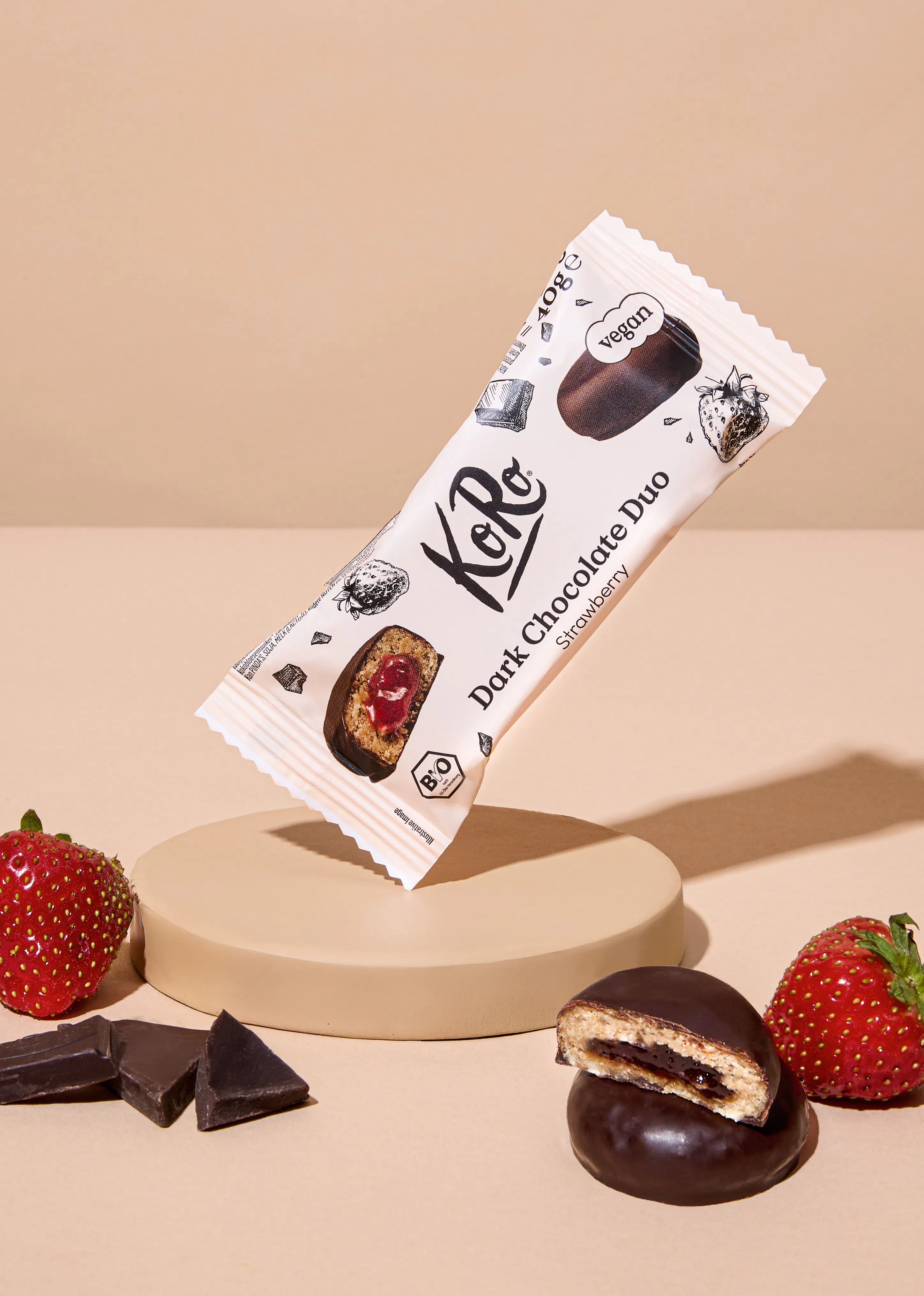 eine schräg schwebende koro dark chocolate duo strawberry riegelpackung, umgeben von frischen erdbeeren, schokoladenstücken und einem aufgeschnittenen riegel, der die erdbeerfüllung zeigt, auf einem beigen podest.