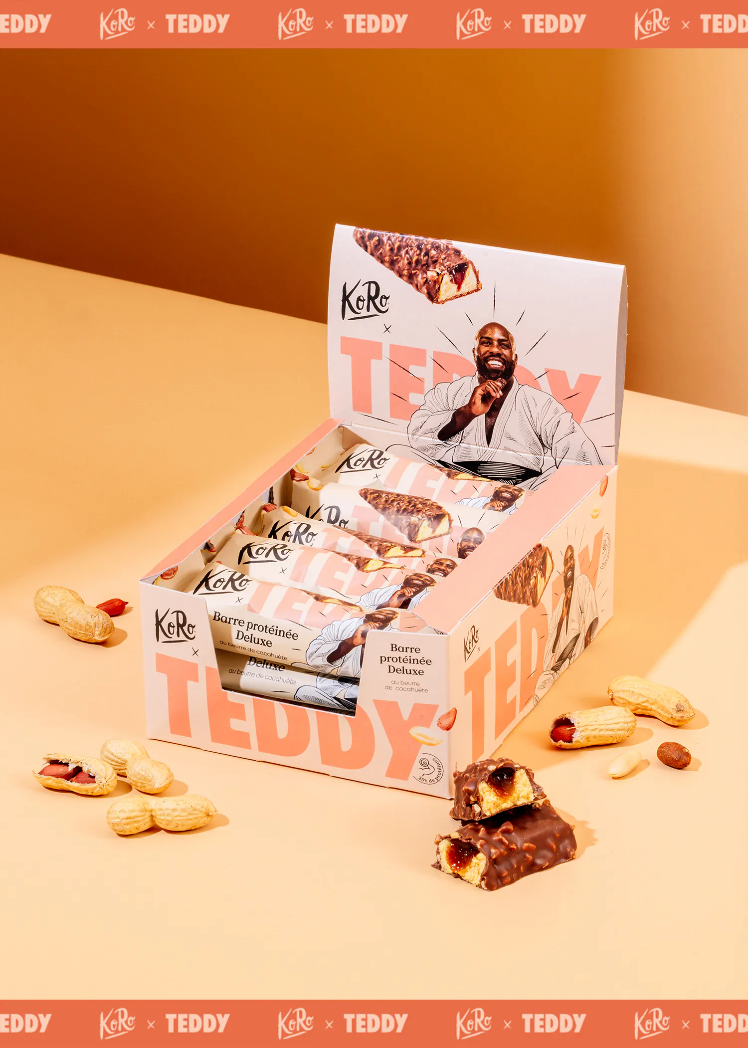 eine offene displaybox mit koro x teddy erdnussbutter-proteinriegeln, umgeben von erdnüssen und zwei aufgeschnittenen riegeln, die die füllung zeigen.