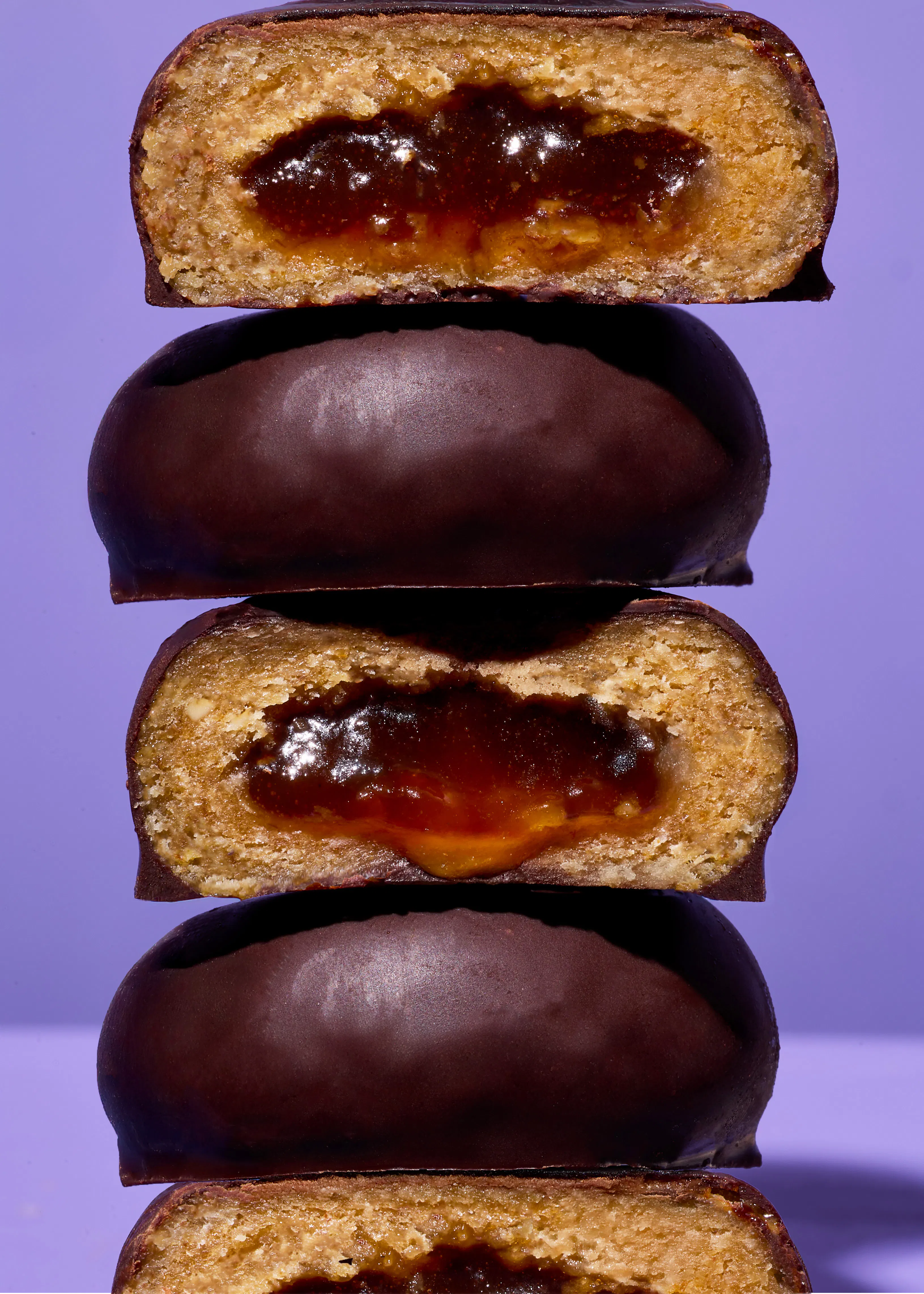 gestapelte schokoladenkekse, von denen einige halbiert sind und eine glänzende fruchtfüllung auf einem lila hintergrund zeigen.