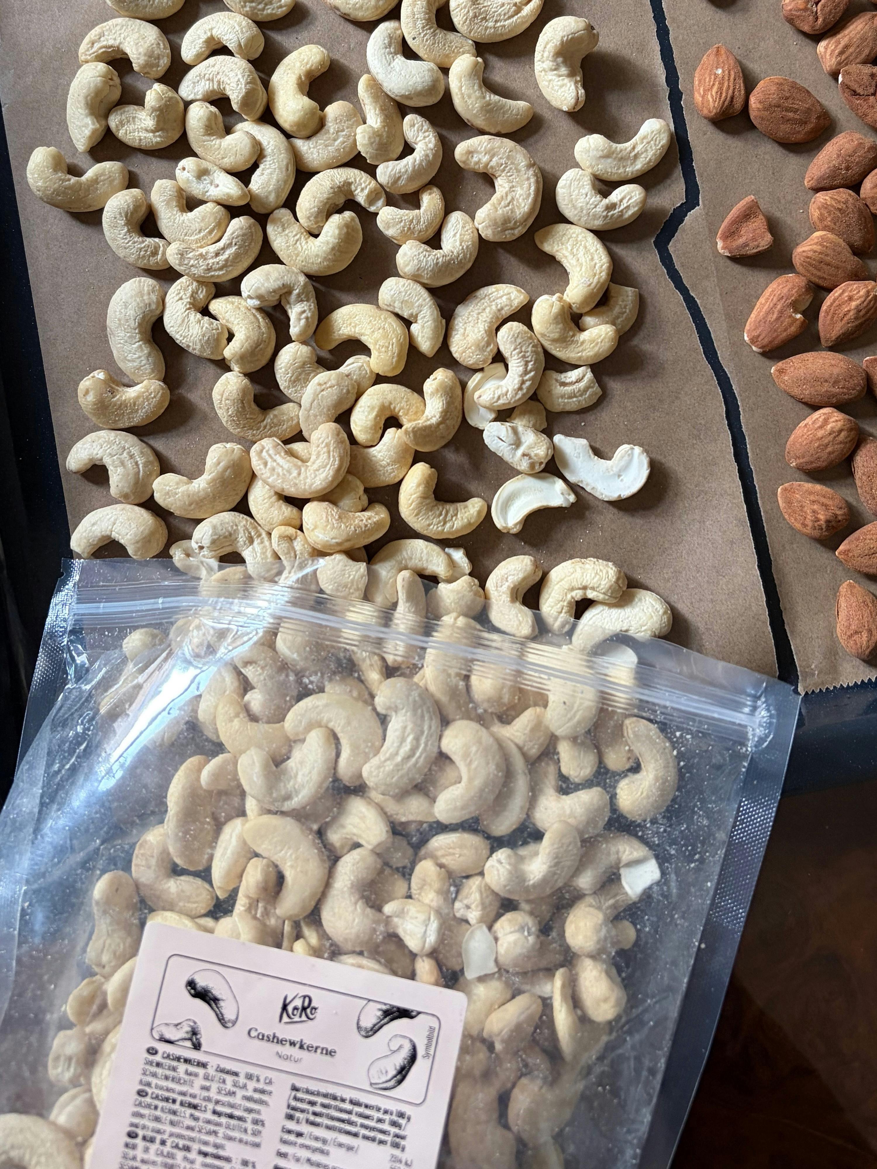 a clear plastic bag of koro cashew nuts sits on a brown paper surface, with some cashews spilled out and a pile of almonds to the right.
