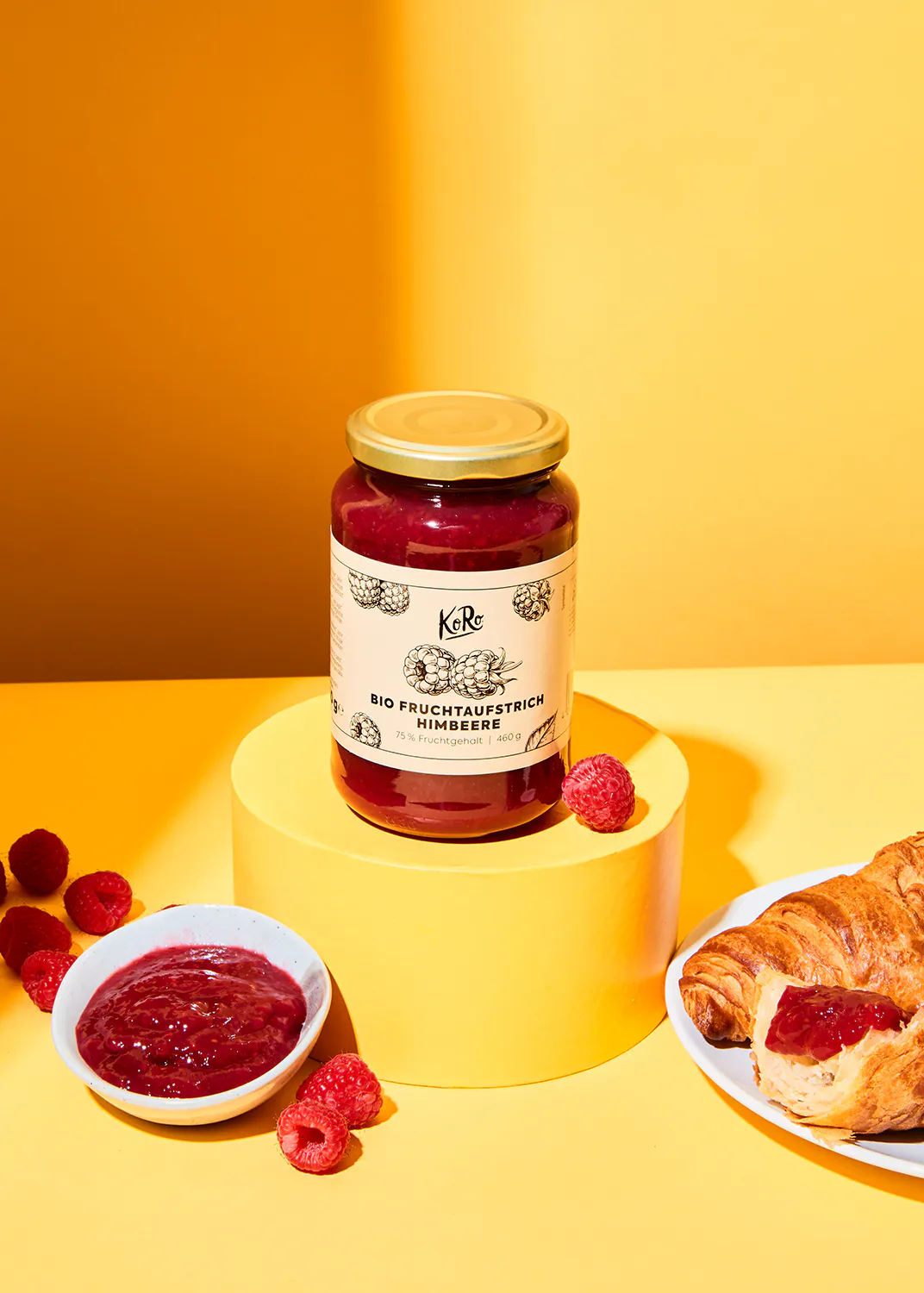 un bocal de confiture de framboises bio est assis sur une table jaune à côté d' un croissant et de framboises