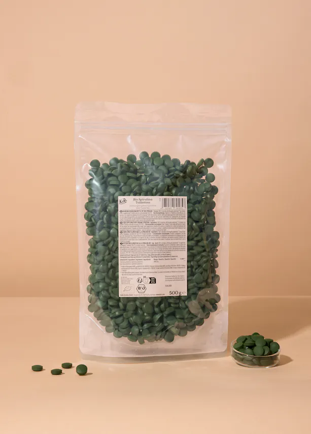 eine transparente plastiktüte mit grünen bio-spirulina-tabletten steht auf einer hellen oberfläche, daneben liegen einige tabletten und eine kleine glasschale mit weiteren tabletten.