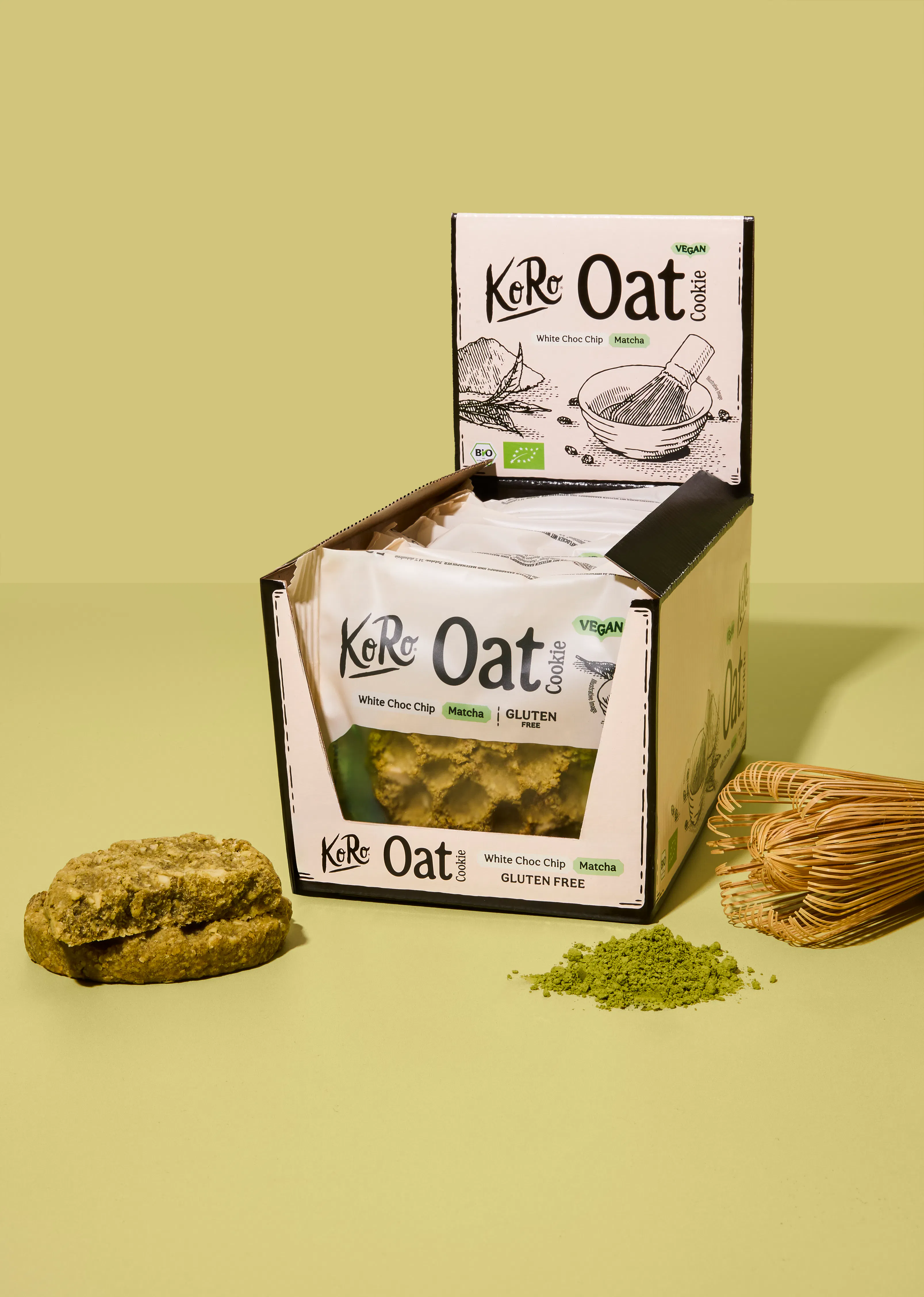 eine verkaufsverpackung mit koro oat cookies in der geschmacksrichtung white choc chip matcha steht neben einem einzelnen keks, etwas matcha-pulver und einem bambusbesen auf einem hellgrünen hintergrund.