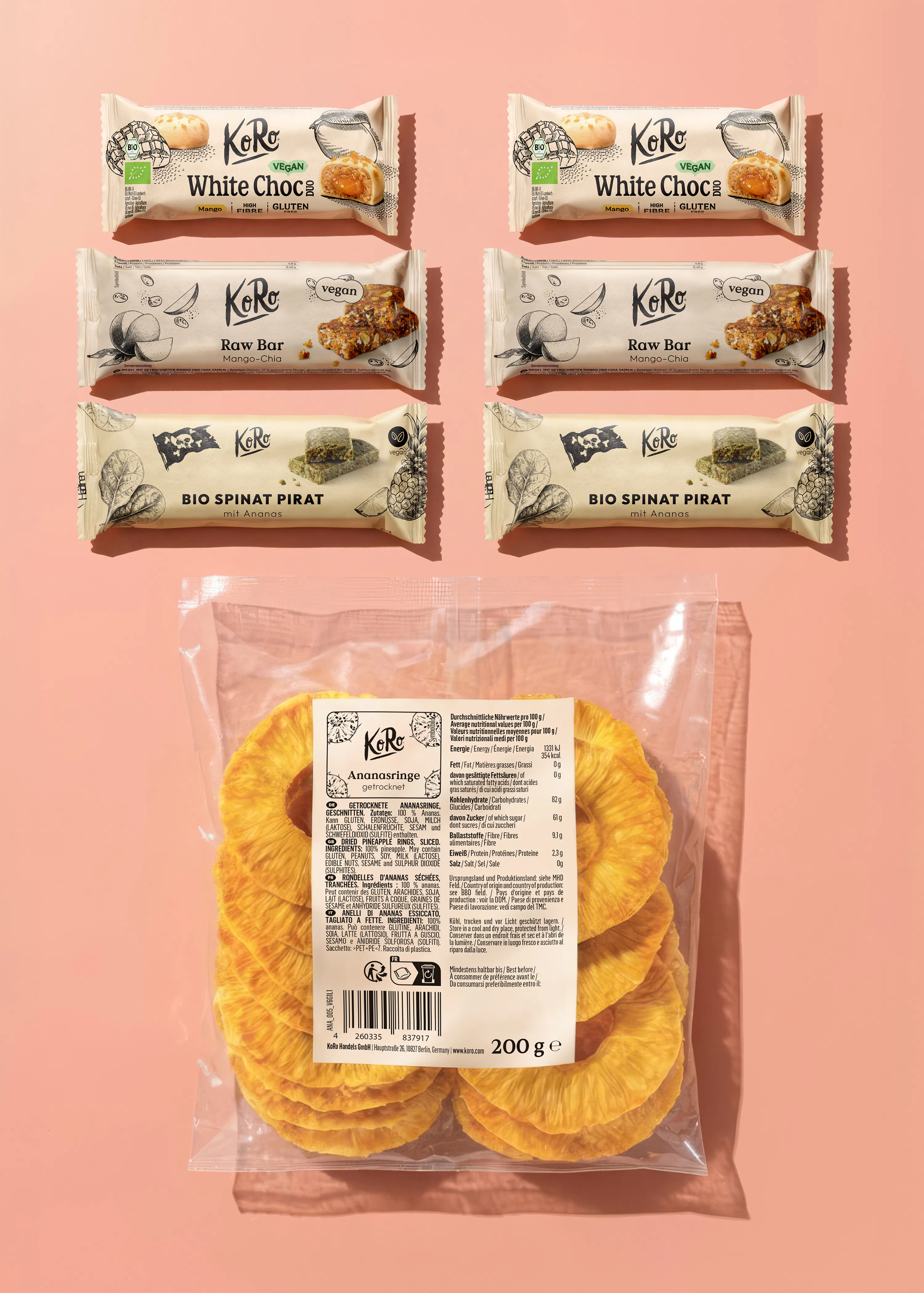 six barres de collation koro (deux mangue chocolat blanc, deux mangue-chia crues, deux épinards ananas bio) et un sachet d'anneaux d'ananas séchés koro sont présentés sur un fond rose.