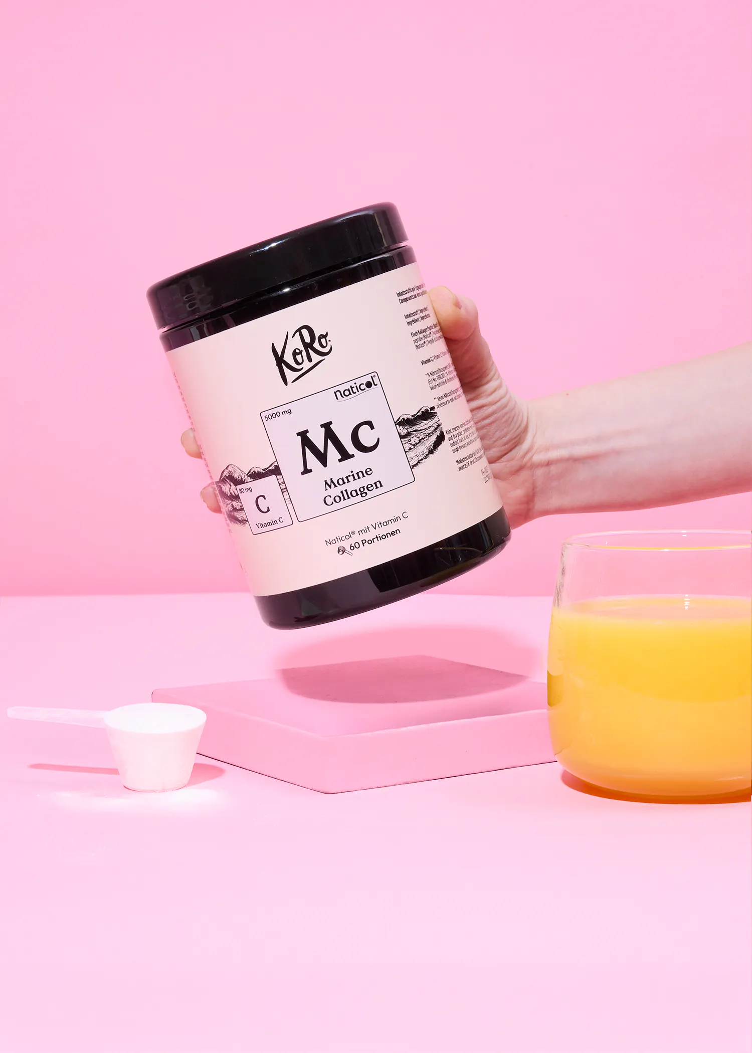 a hand holds a white and black container of koro naticol marine collagen powder, with a scoop of powder and a glass of orange juice on a pink surface.