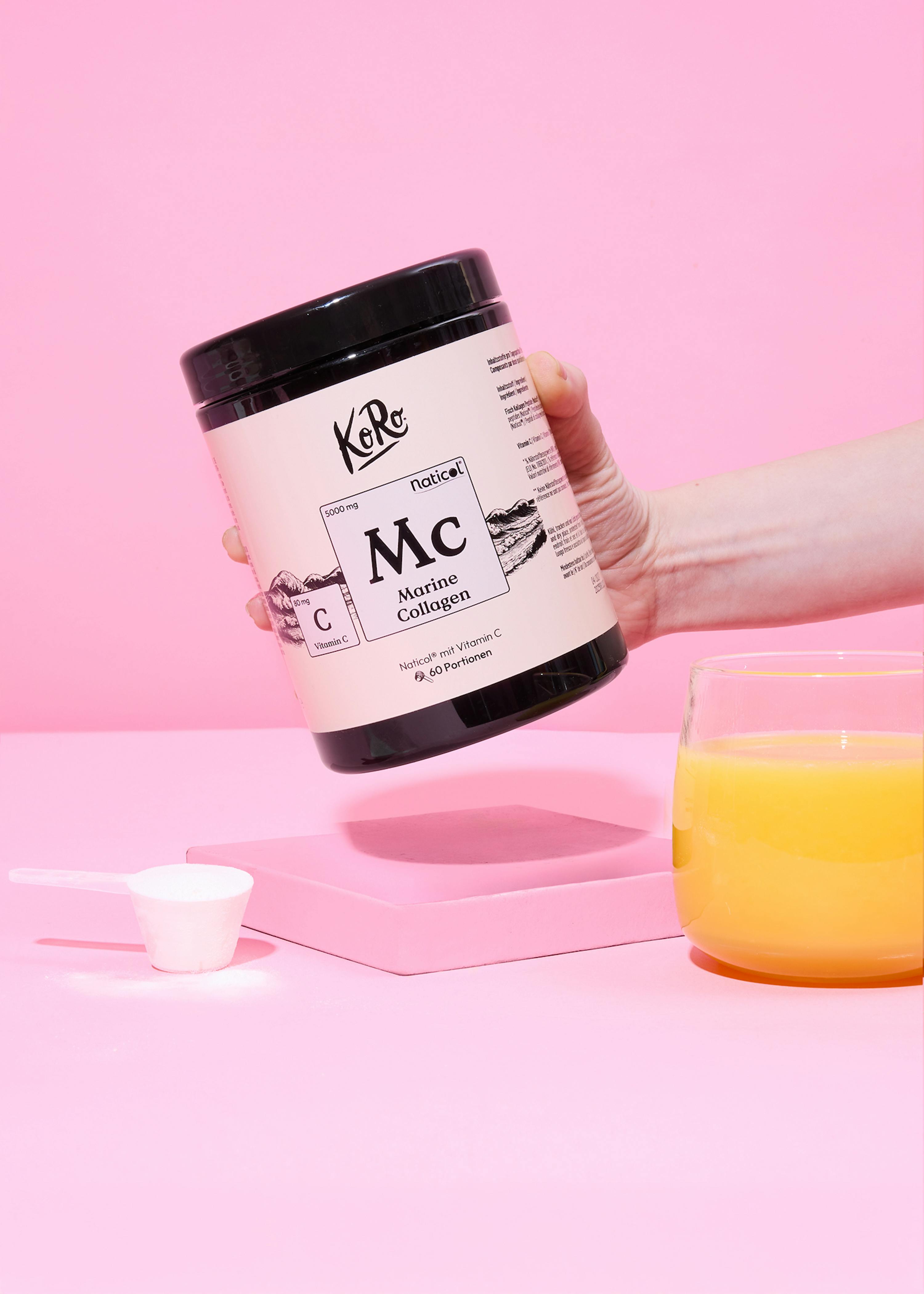 eine hand hält einen beigefarbenen behälter mit der aufschrift "koro naticol mc marine collagen" und "vitamin c", daneben stehen ein weißer messlöffel mit pulver und ein glas orangensaft auf einer pinken oberfläche.