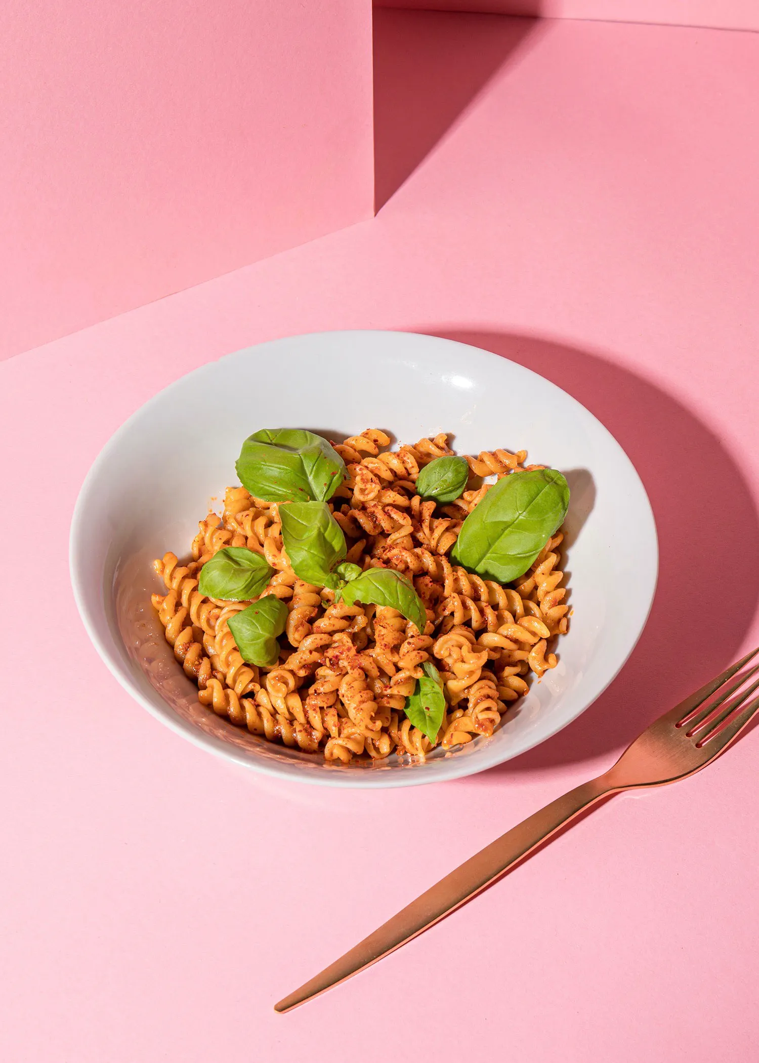 un bol de pâtes avec des feuilles de basilic et une fourchette sur une table rose