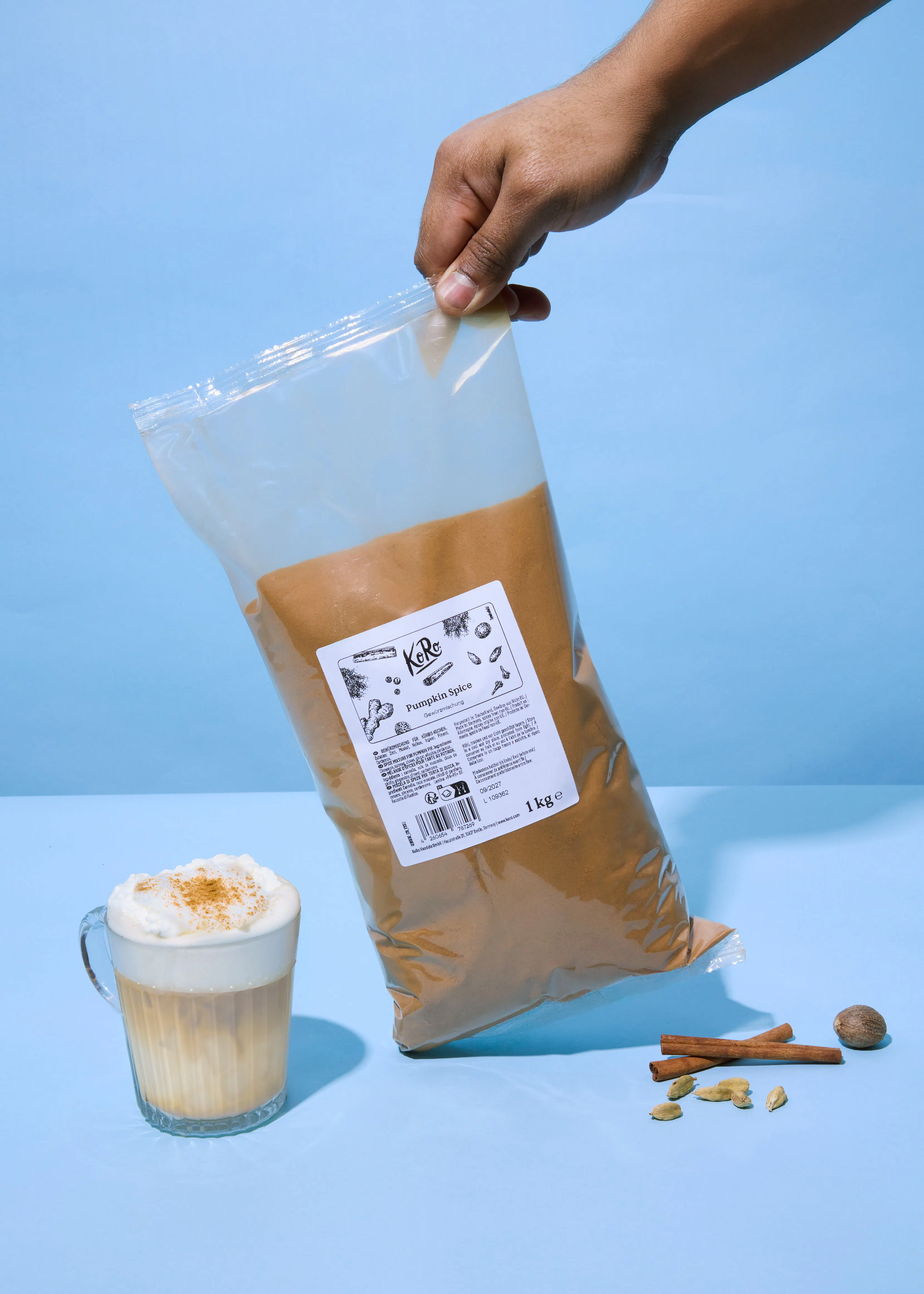 une main tient un grand sac d'épices à citrouille au-dessus d'une boisson avec de la crème fouettée et d'épices entières (cannelle, cardamome, muscade) sur un fond bleu.