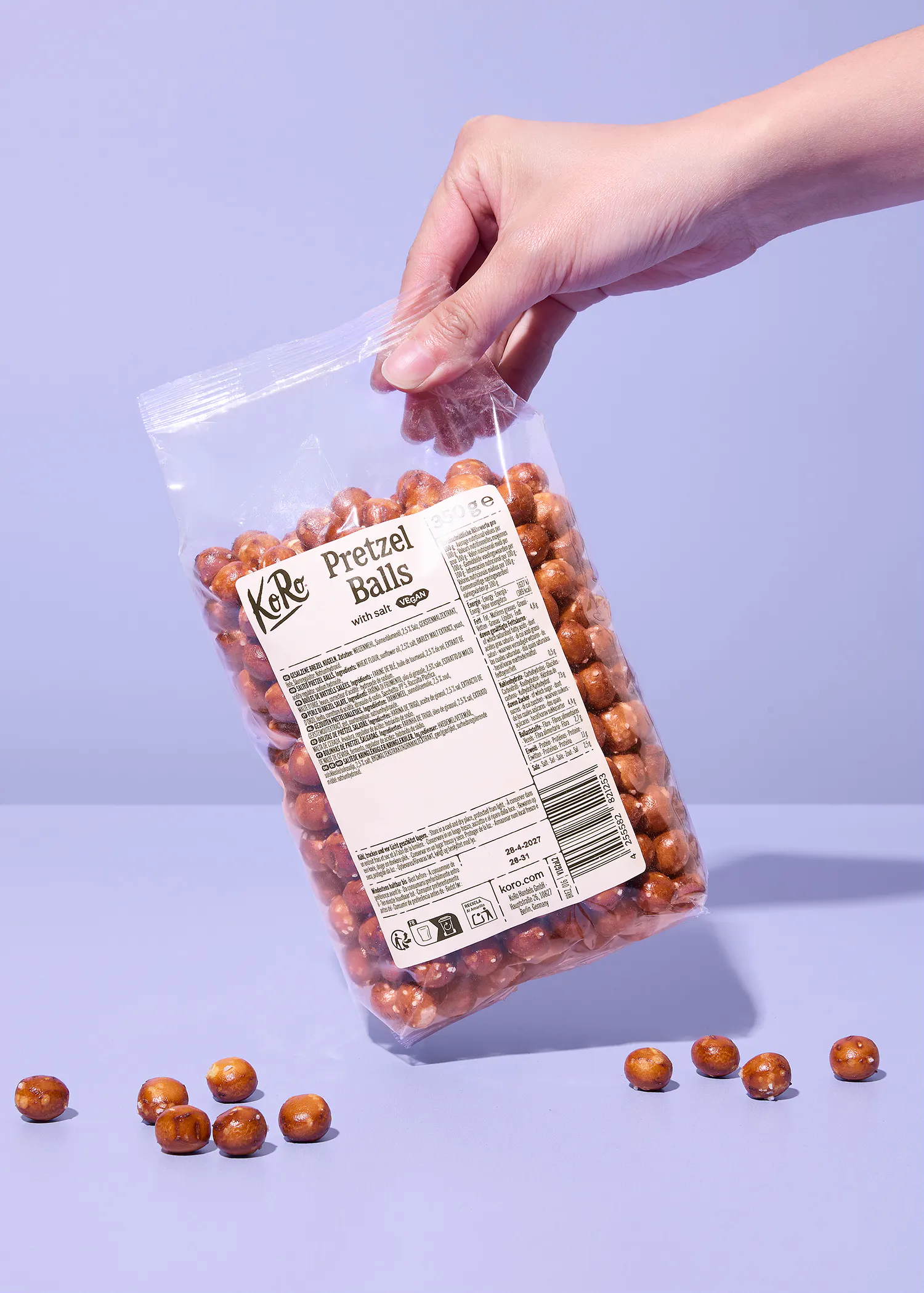 eine hand hält eine durchsichtige tüte mit koro pretzel balls vor einem hellvioletten hintergrund, wobei einige der salzigen teigkugeln auf der fläche darunter verstreut liegen.