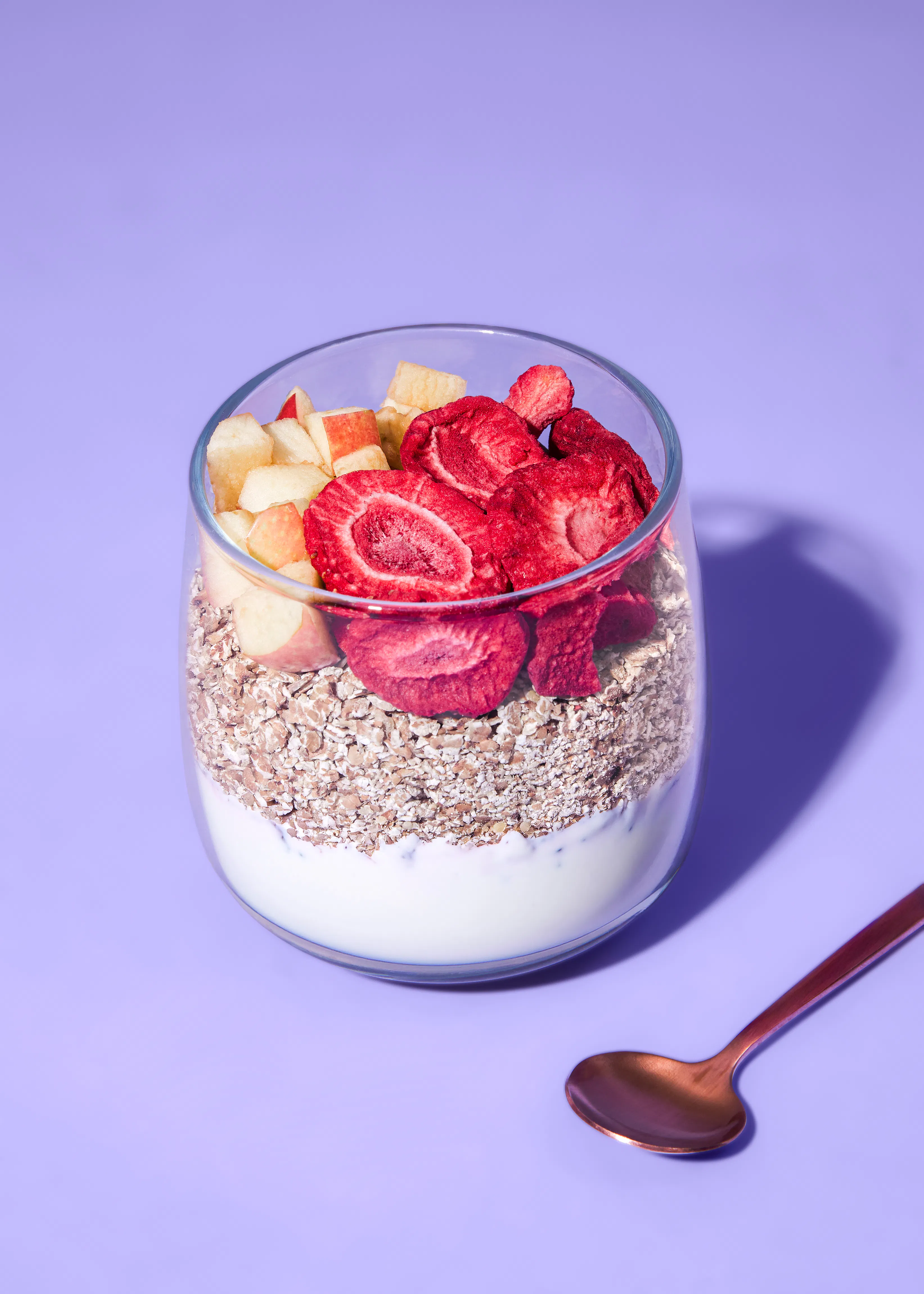 un vaso de cristal con capas de yogur, avena, trozos de manzana y fresas liofilizadas, junto a una cuchara de cobre sobre un fondo morado.