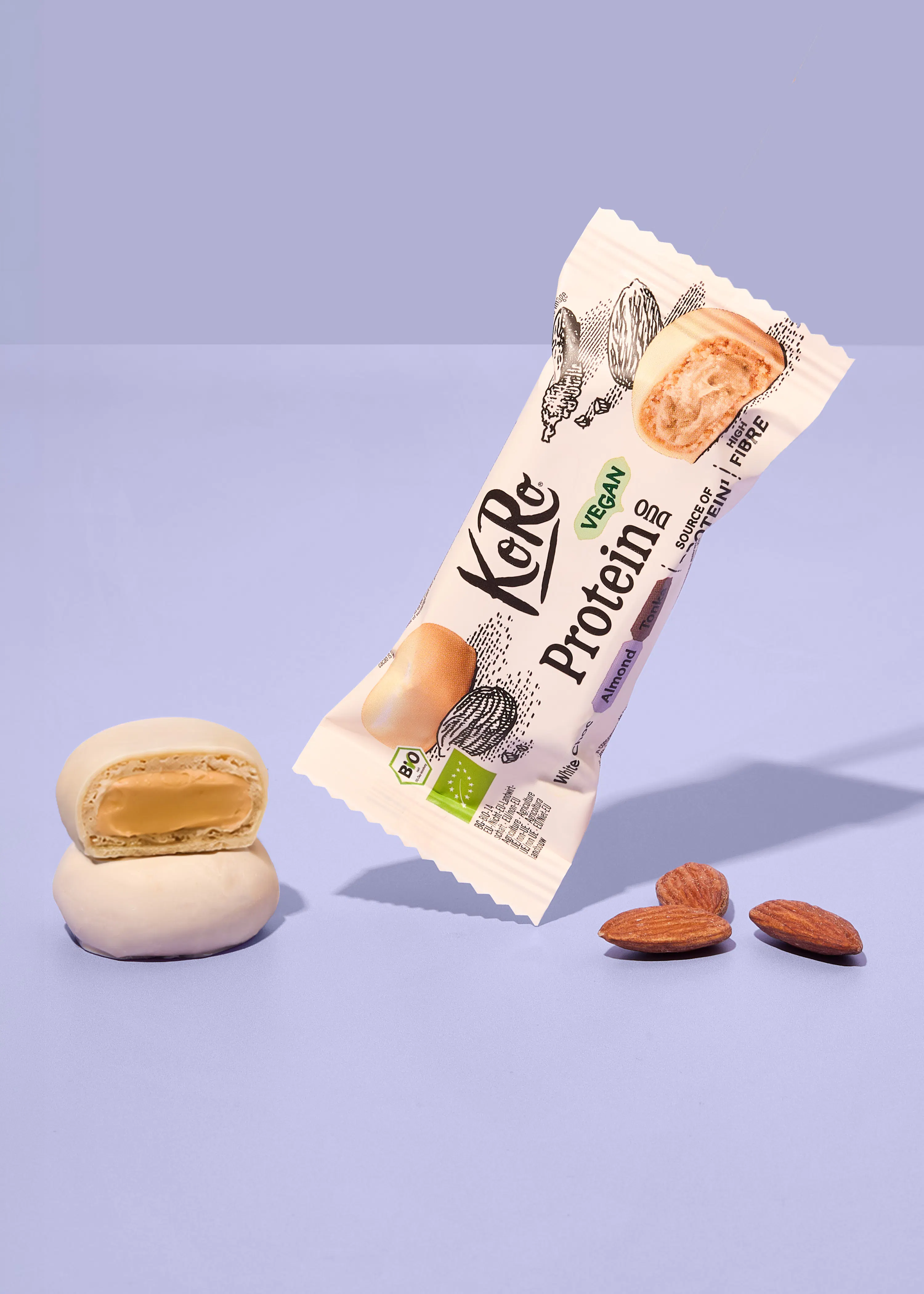 ein koro proteinriegel in weißer schokolade mit mandeln liegt schräg neben zwei halbierten riegeln und drei ganzen mandeln auf einem hellvioletten hintergrund.