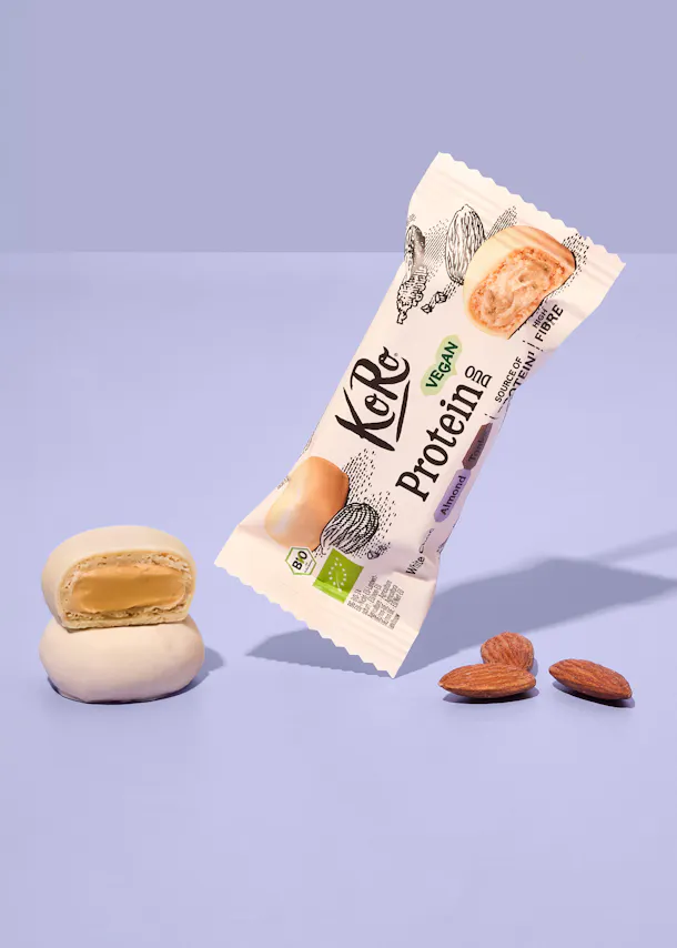 ein koro proteinriegel in weißer schokolade mit mandeln liegt schräg neben zwei halbierten riegeln und drei ganzen mandeln auf einem hellvioletten hintergrund.