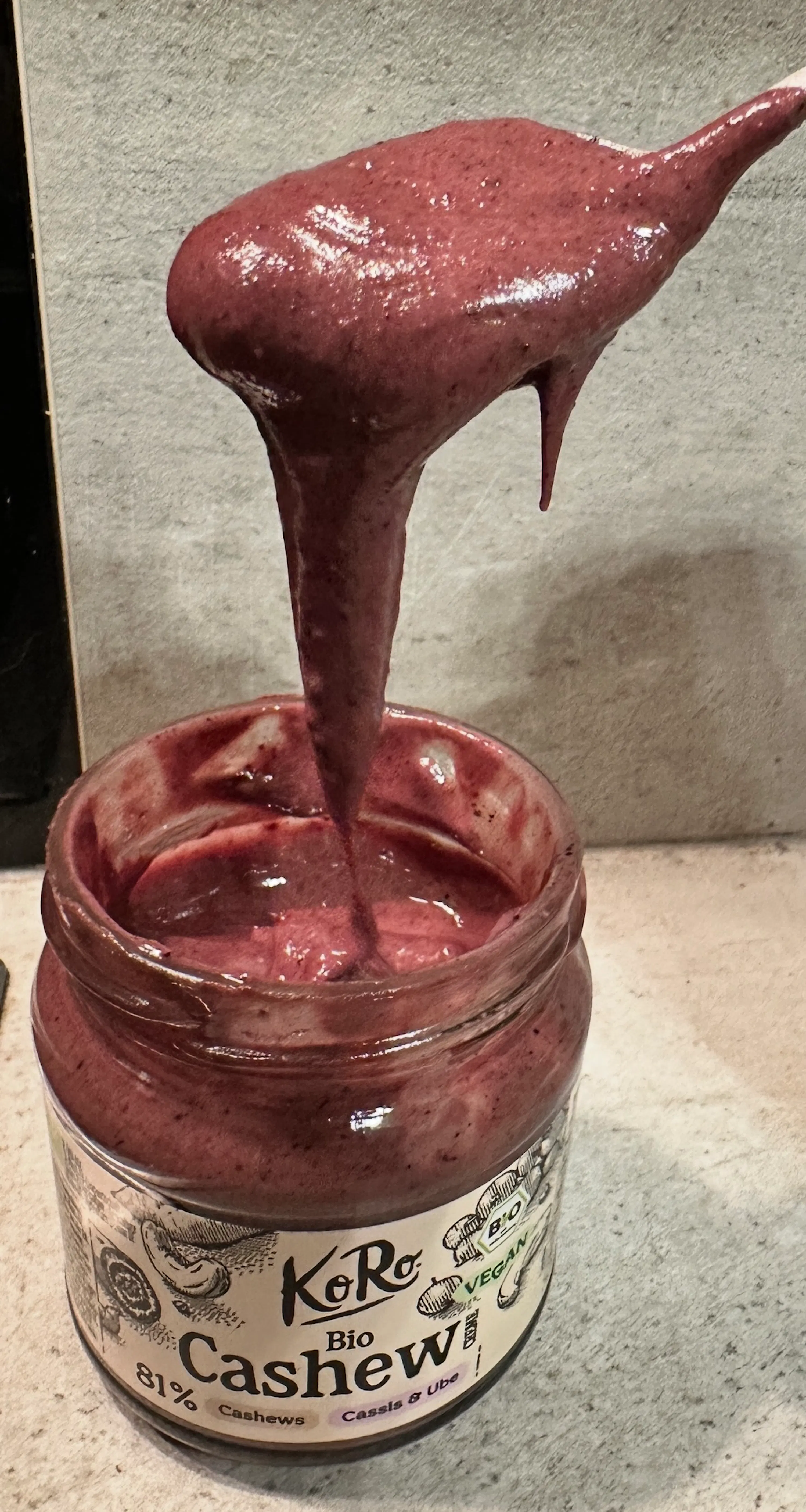 a spoon lifts thick, purple-red cashew spread from a glass jar labeled "koro bio cashew cassis & ube", with some spread dripping back into the jar.