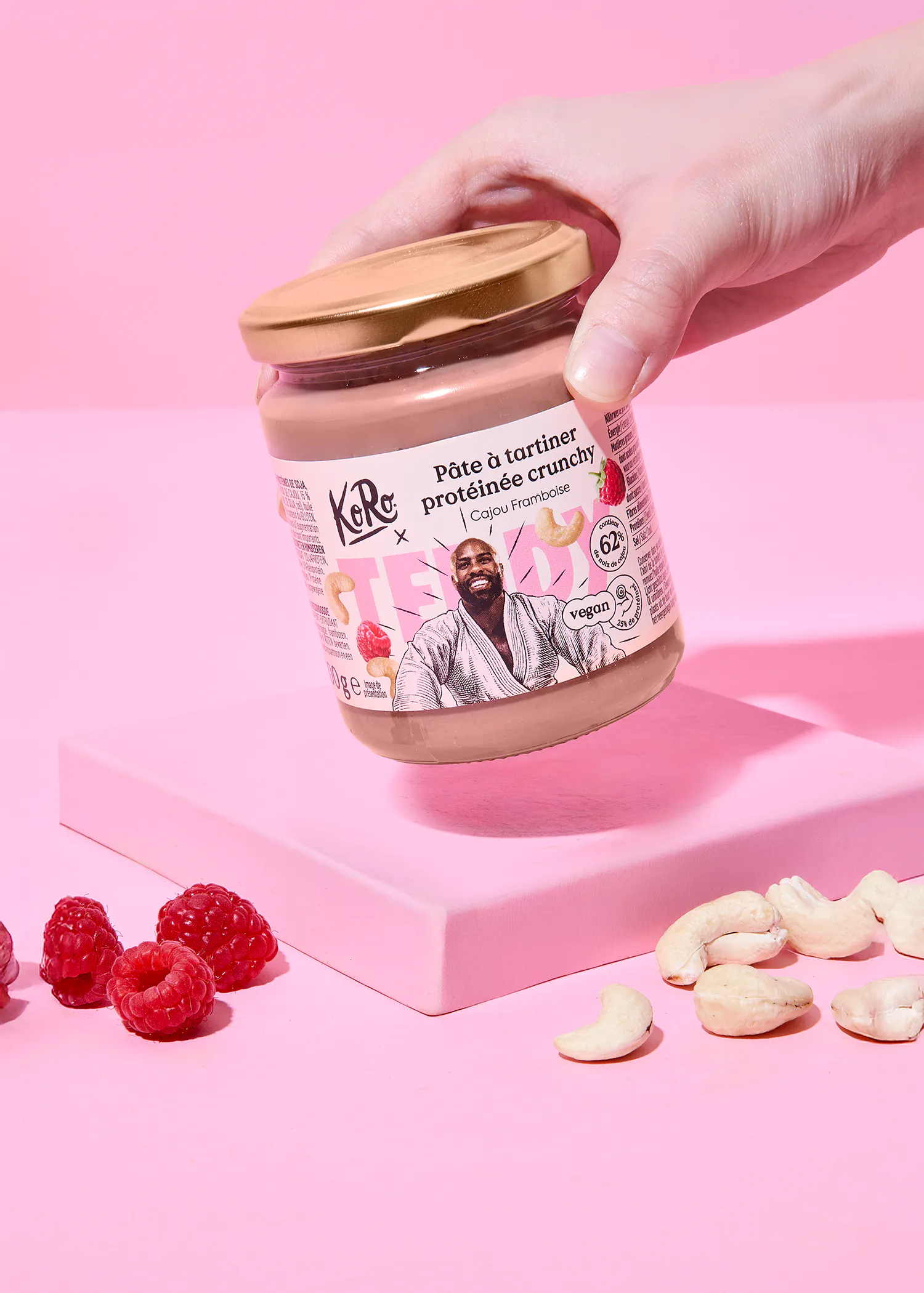 une main tient un pot de pâte à tartiner protéinée crunchy cajou framboise koro au-dessus d'une surface rose avec des framboises et des noix de cajou éparpillées.