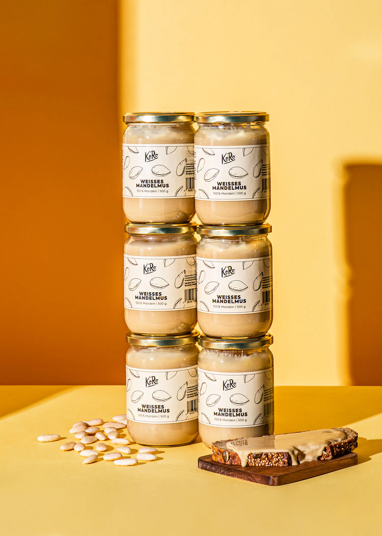 six pots de beurre d'amande blanc koro sont empilés sur un fond jaune, avec des amandes éparpillées et une tranche de pain recouverte de beurre d'amande sur une planche en bois.
