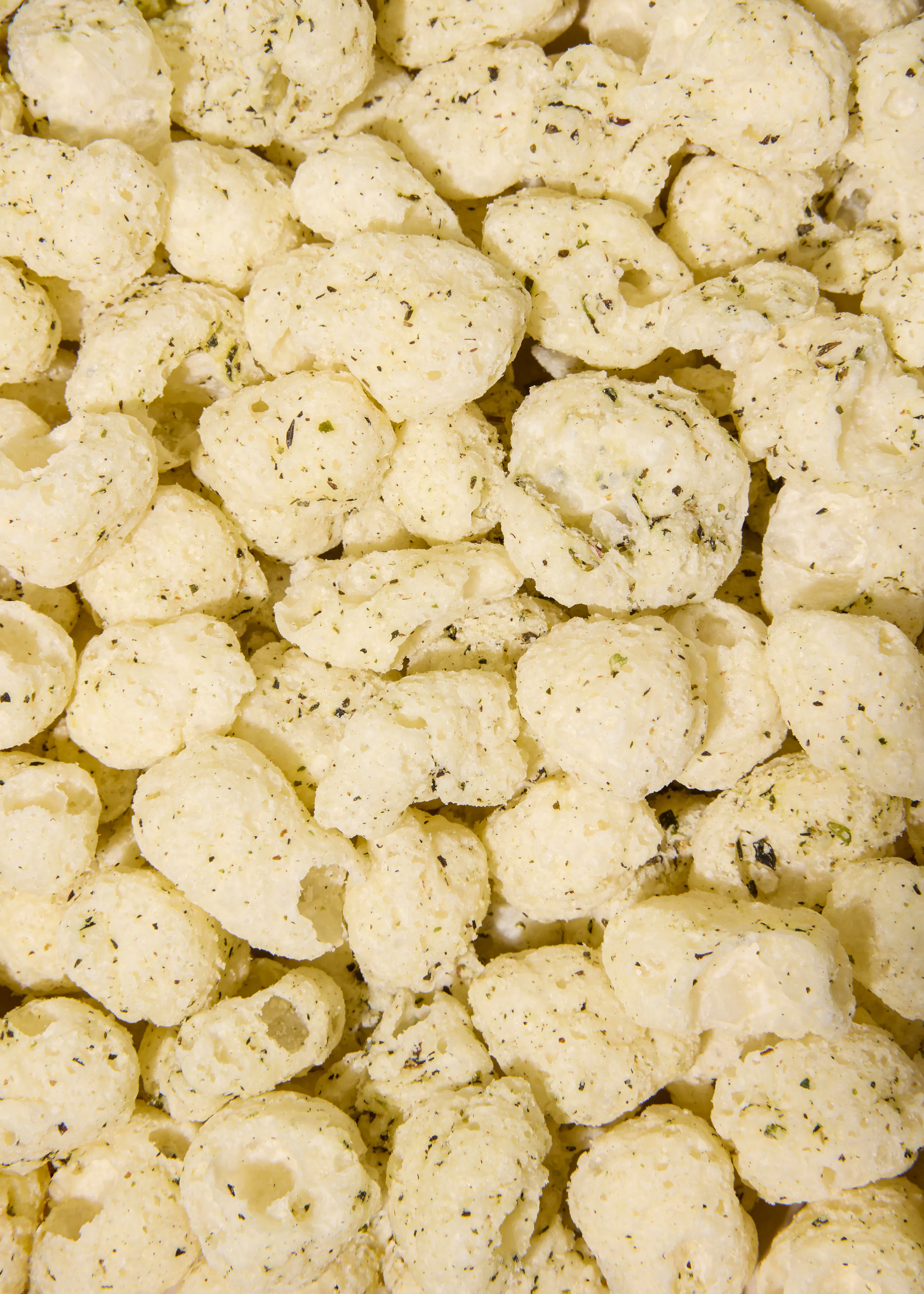 a close-up view of a pile of light-colored, puffed corn snacks seasoned with small green herbs.