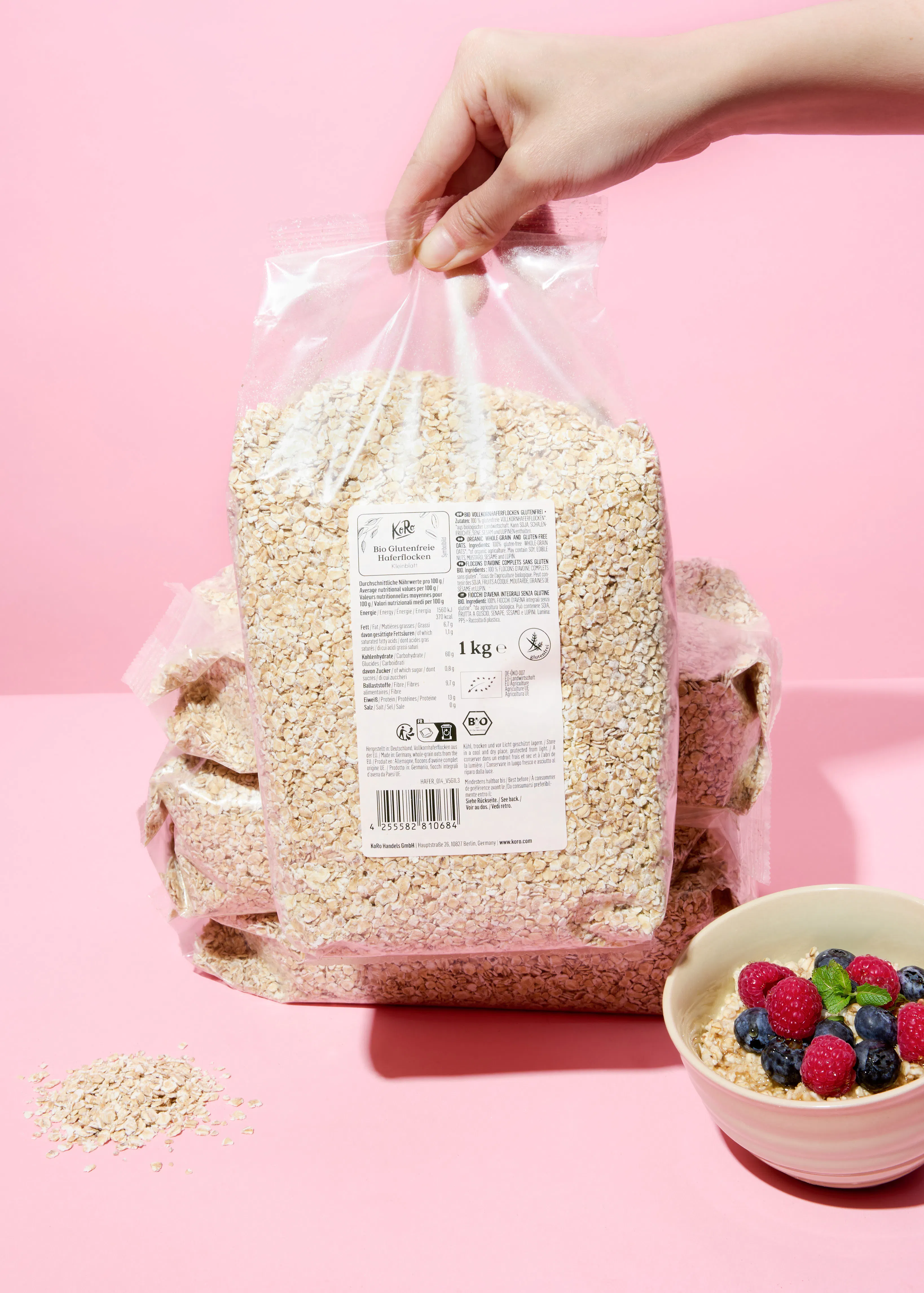 eine hand hält eine transparente tüte mit bio glutenfreien haferflocken, daneben steht eine schale haferbrei mit beeren auf einem pinken hintergrund.
