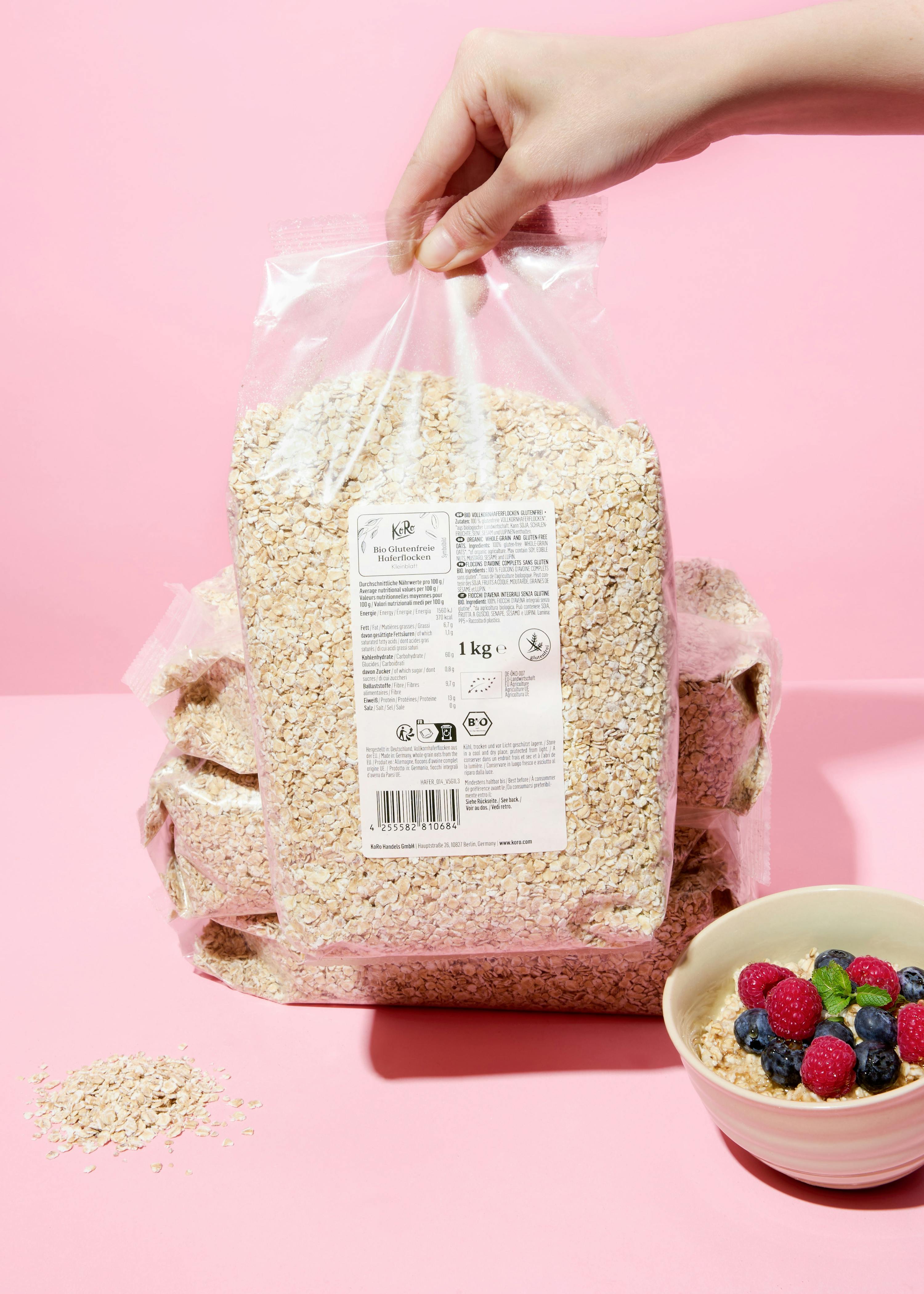 a hand holds a 1kg bag of gluten-free oats, with a bowl of oatmeal topped with berries and mint, and loose oats on a pink surface.