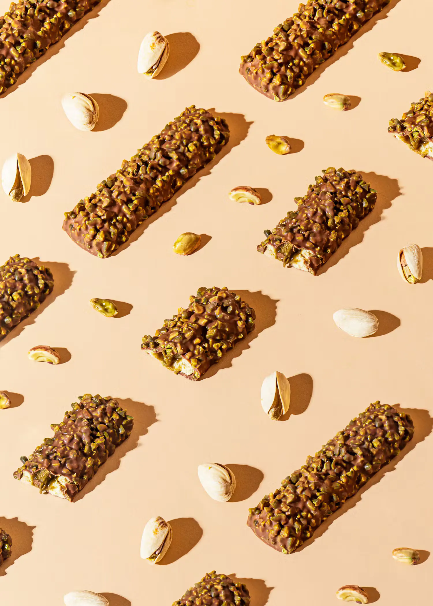 plusieurs barres chocolatées garnies de pistaches concassées, accompagnées de pistaches entières et décortiquées, sur un fond beige.
