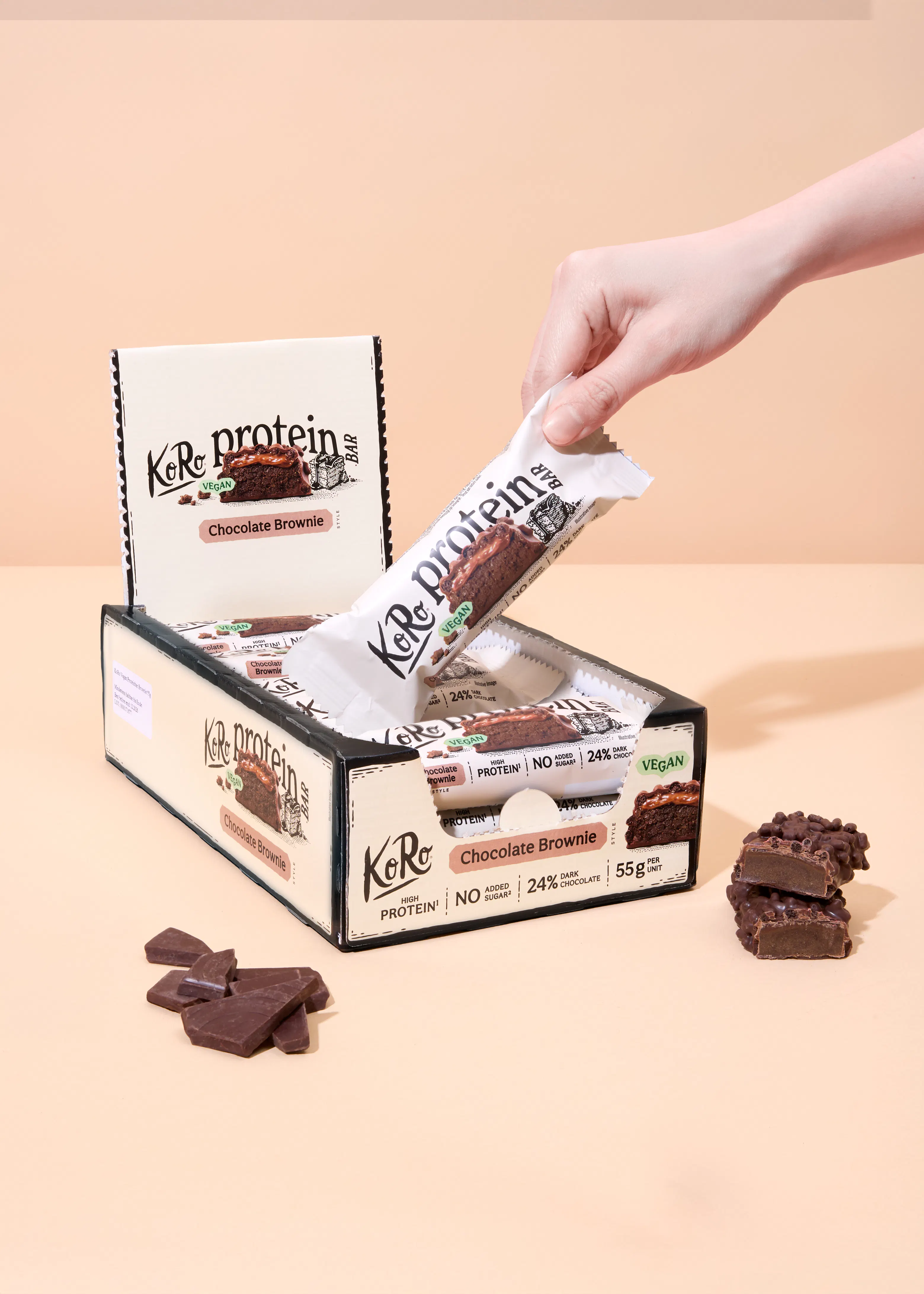 a hand removes a chocolate brownie flavored koro protein bar from an open display box, with loose chocolate pieces and a cut bar visible nearby.