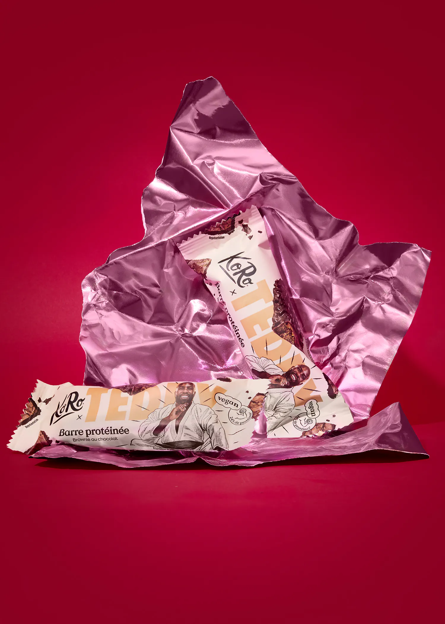 deux barres protéinées koro saveur brownie au chocolat, avec un emballage illustré d'un homme en kimono et la mention "vegan", sont posées sur un papier d'aluminium froissé rose sur un fond rouge.