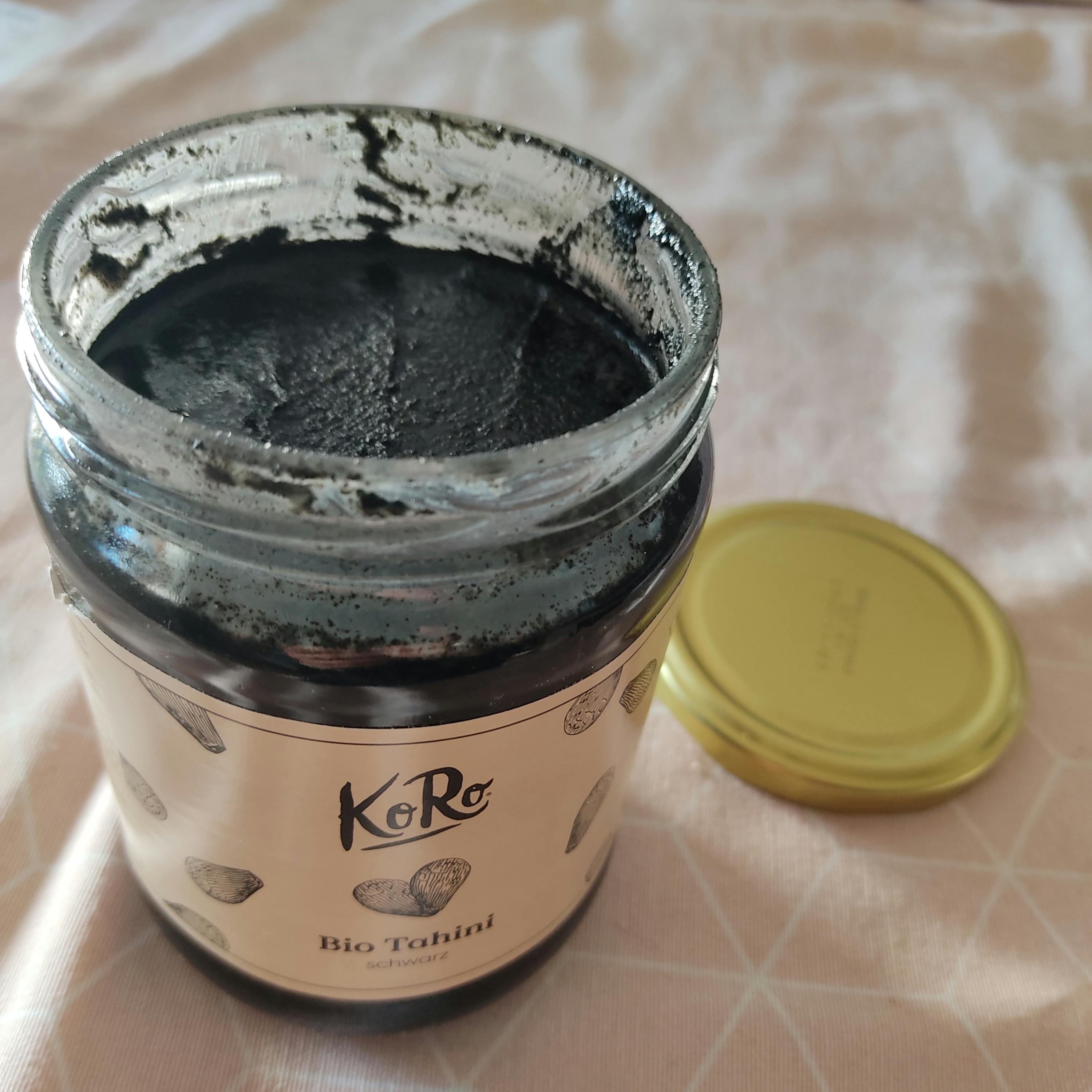 un pot en verre ouvert de tahini noir de la marque koro, avec son couvercle doré posé à côté sur un tissu clair.