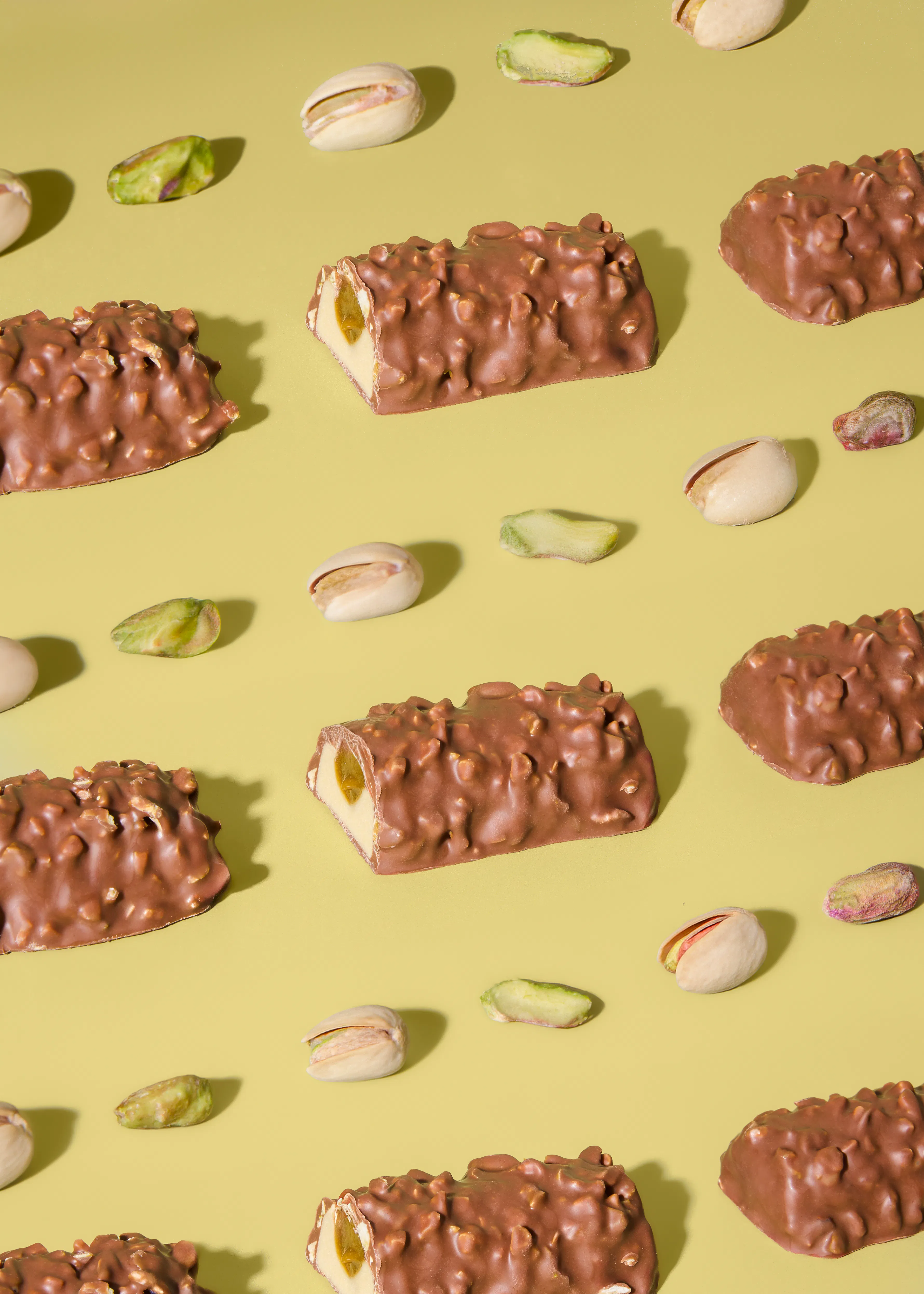 una composizione di barrette di cioccolato con noci, alcune tagliate per mostrare un ripieno cremoso al pistacchio, e pistacchi sparsi su uno sfondo giallo chiaro.