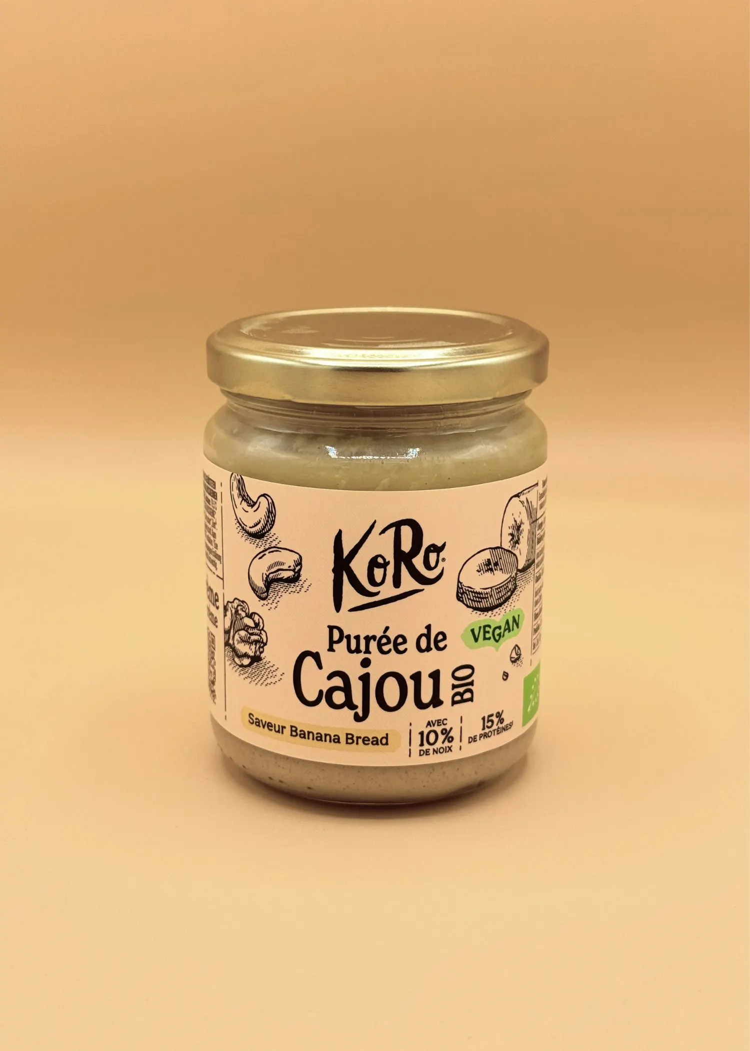 ein glas koro bio-cashewmus mit bananenbrotgeschmack, vegan, mit 10% nüssen und 15% protein, steht auf einem hellbraunen hintergrund.