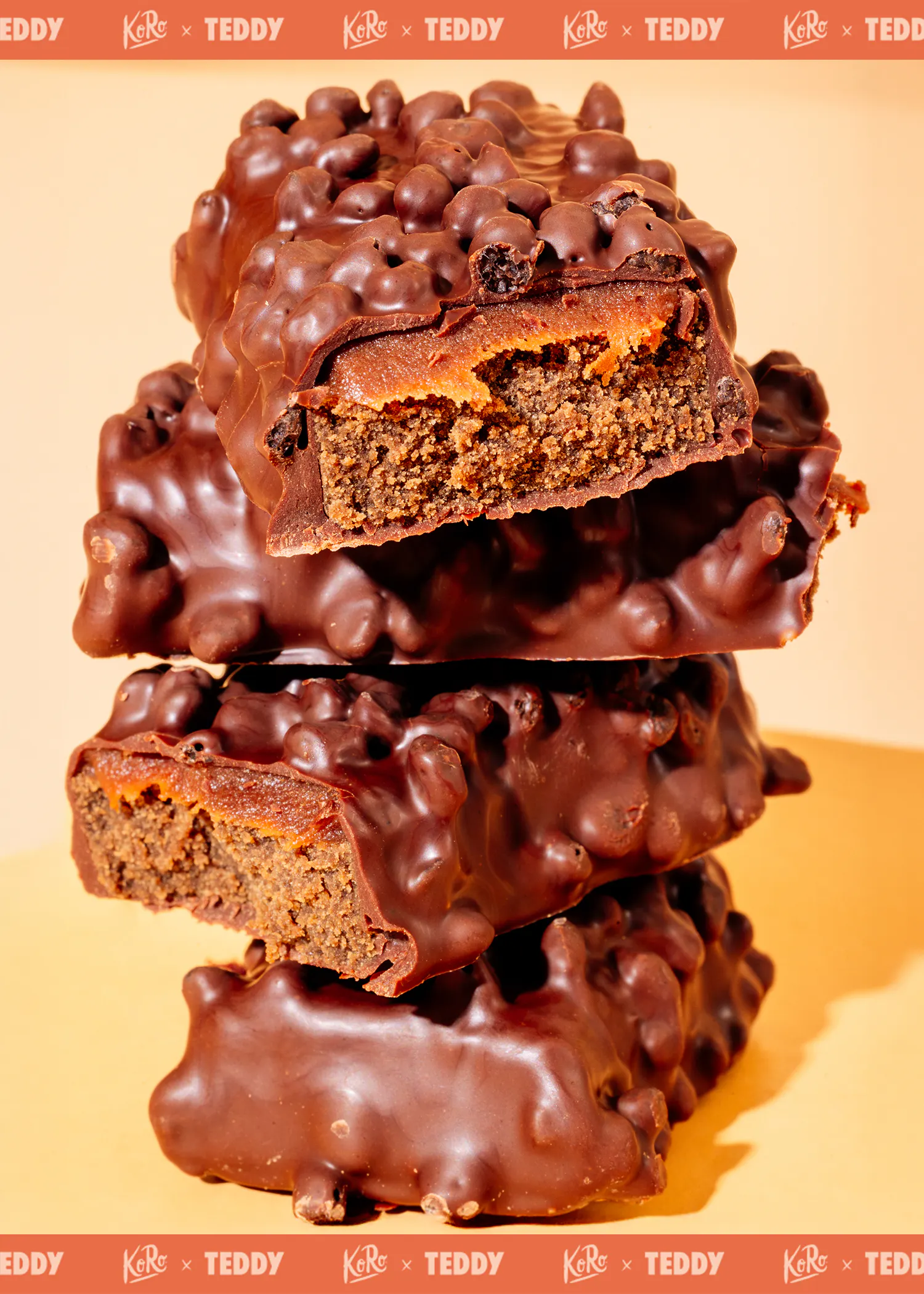 trois barres chocolatées empilées, la barre du dessus étant cassée pour révéler un intérieur de gâteau brun foncé et une garniture orange sous un enrobage de chocolat croustillant.