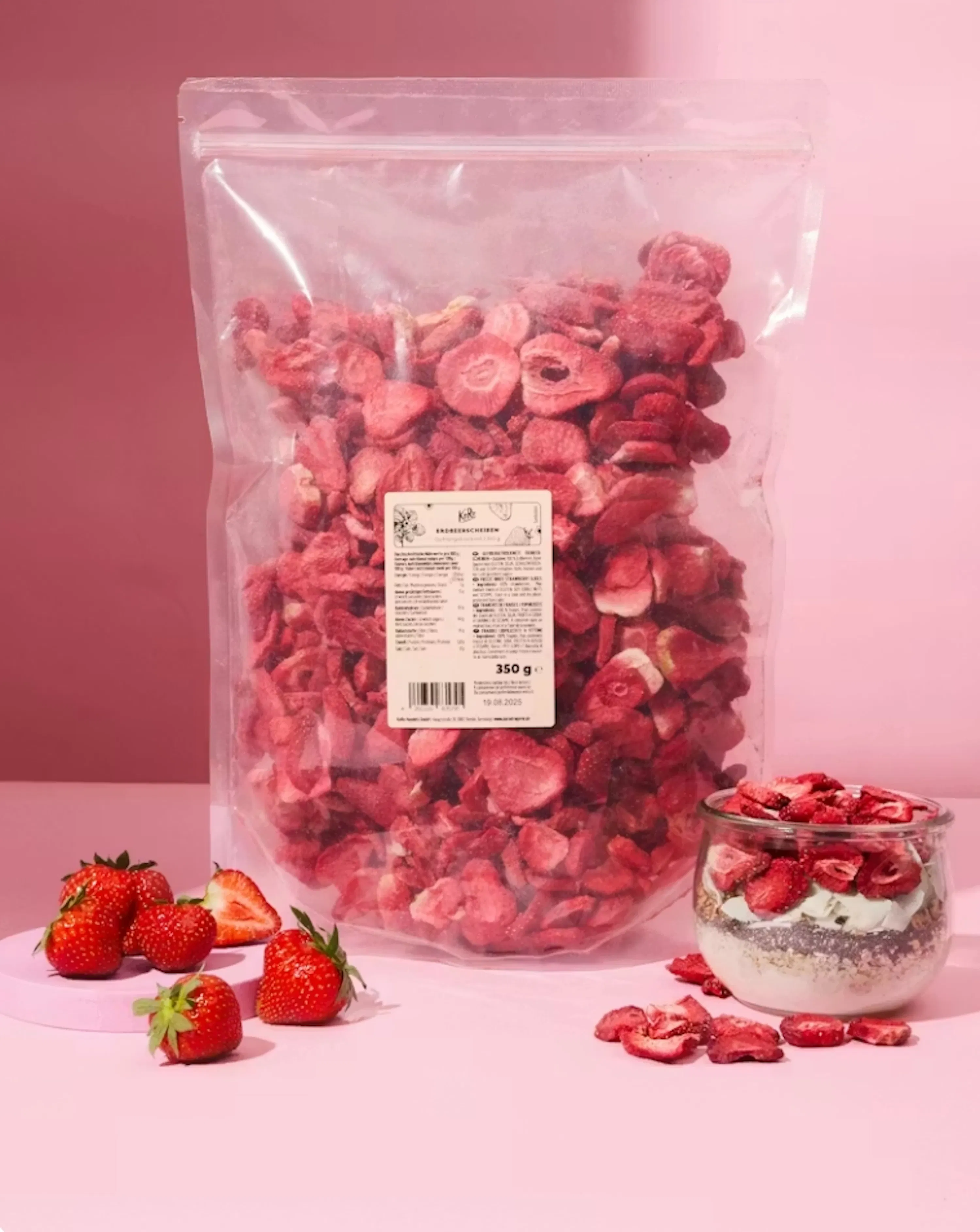 ein großer transparenter beutel mit gefriergetrockneten erdbeerscheiben steht neben frischen erdbeeren und einem glas mit joghurt und gefriergetrockneten erdbeeren auf einem rosa hintergrund.