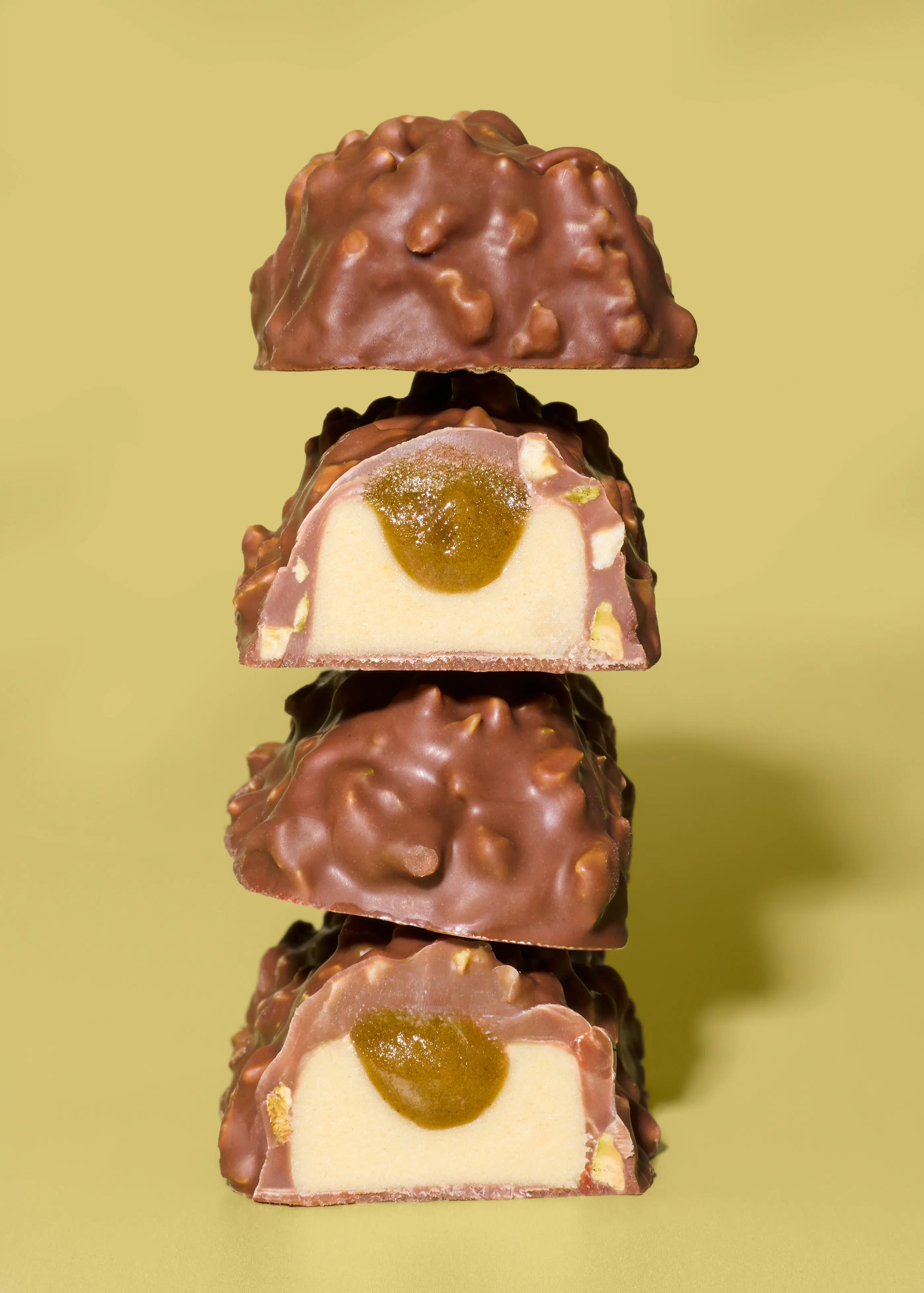 ein stapel aus vier schokoladenüberzogenen eisriegeln mit nussstückchen, wobei zwei aufgeschnitten sind und eine cremige füllung mit einem grünen kern zeigen, vor einem hellgrünen hintergrund.