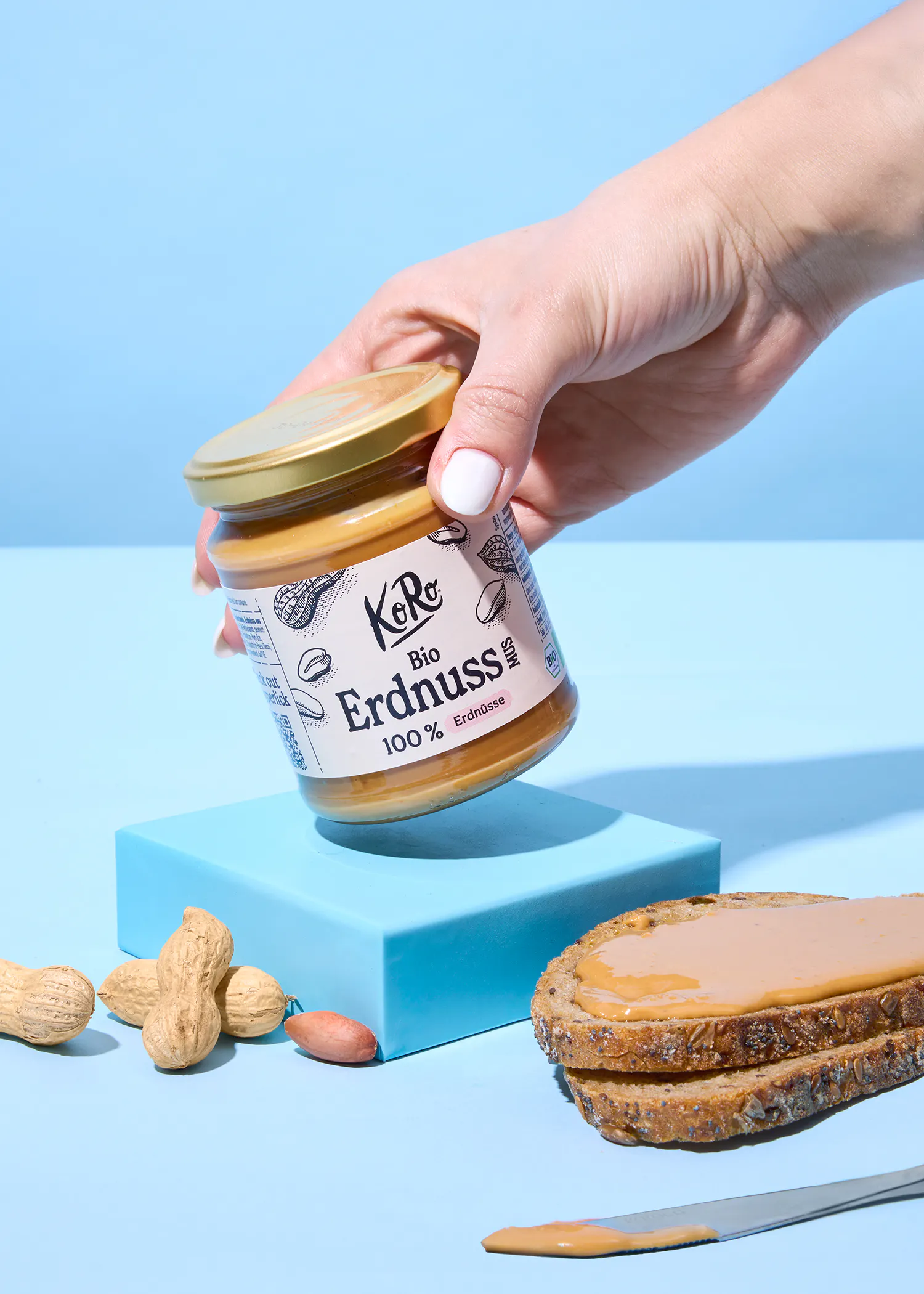 une main tient un pot de beurre de cacahuète koro bio erdnuss au-dessus d'une tranche de pain grillé tartinée, d'un couteau et de cacahuètes sur un fond bleu.