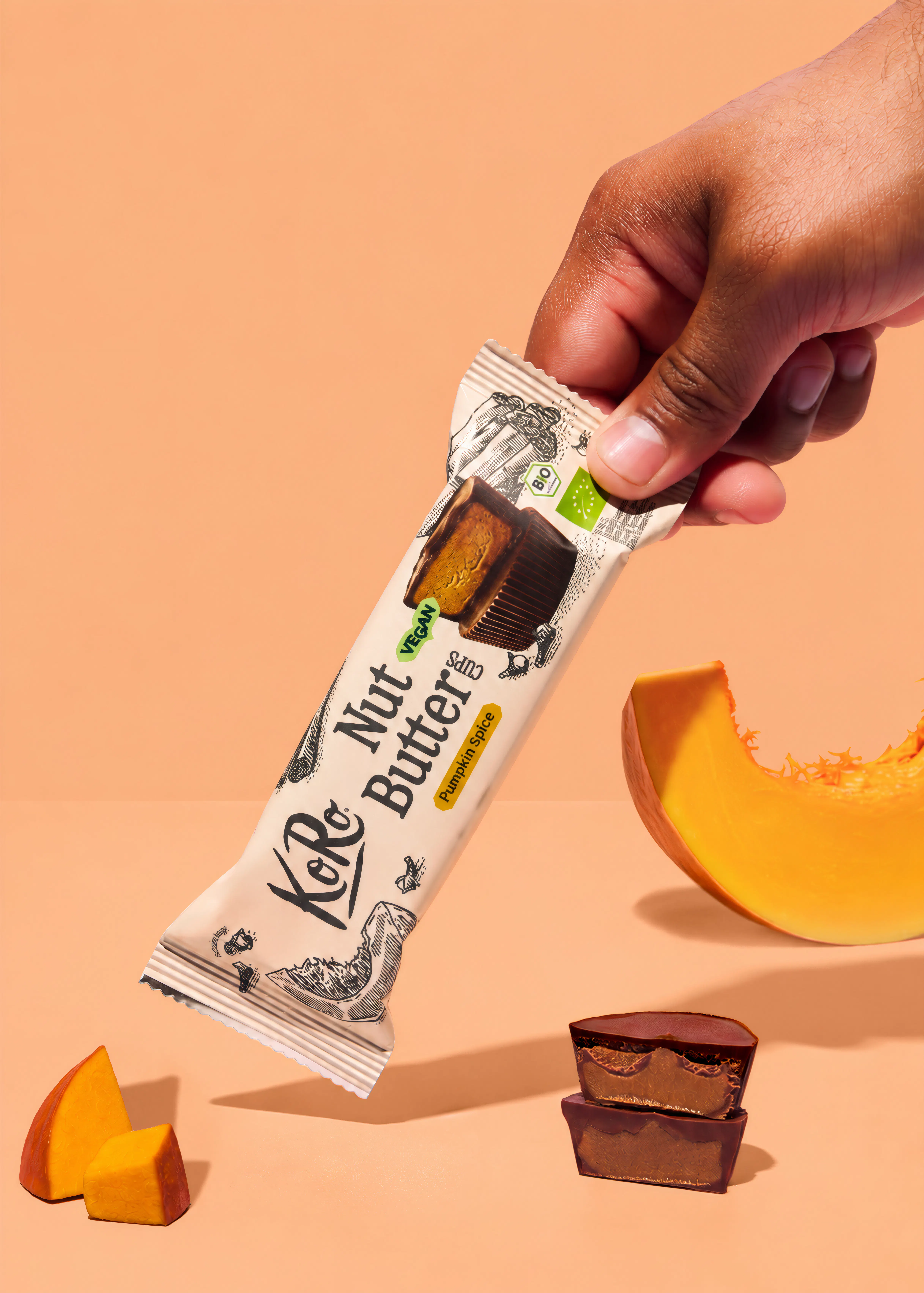 una mano tiene una confezione di koro nut butter cups al gusto pumpkin spice accanto a pezzi di zucca e due cioccolatini aperti su sfondo color pesca.