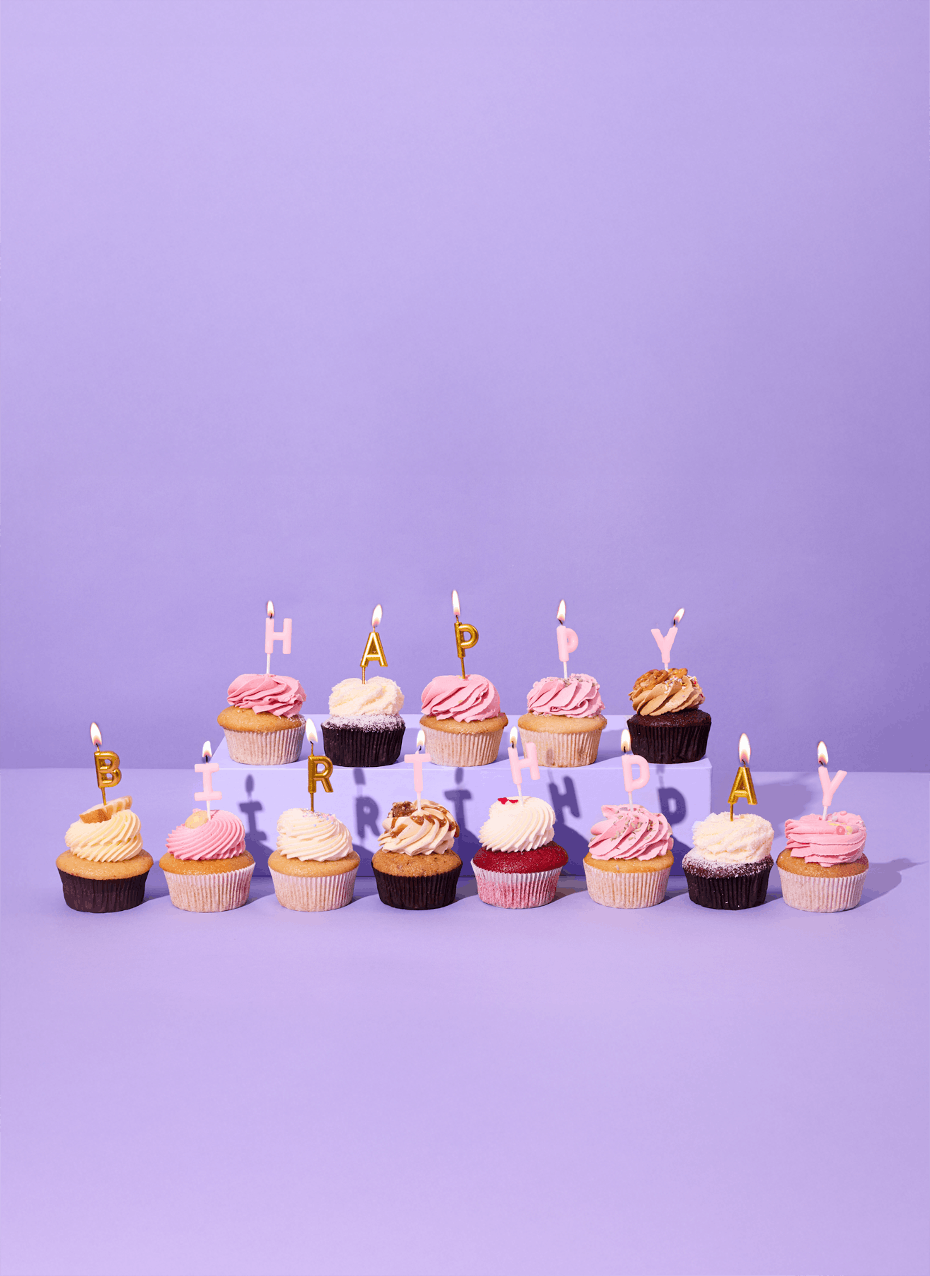 douze cupcakes colorés avec des bougies allumées formant les mots 'happy birthday' sur un fond lavande.