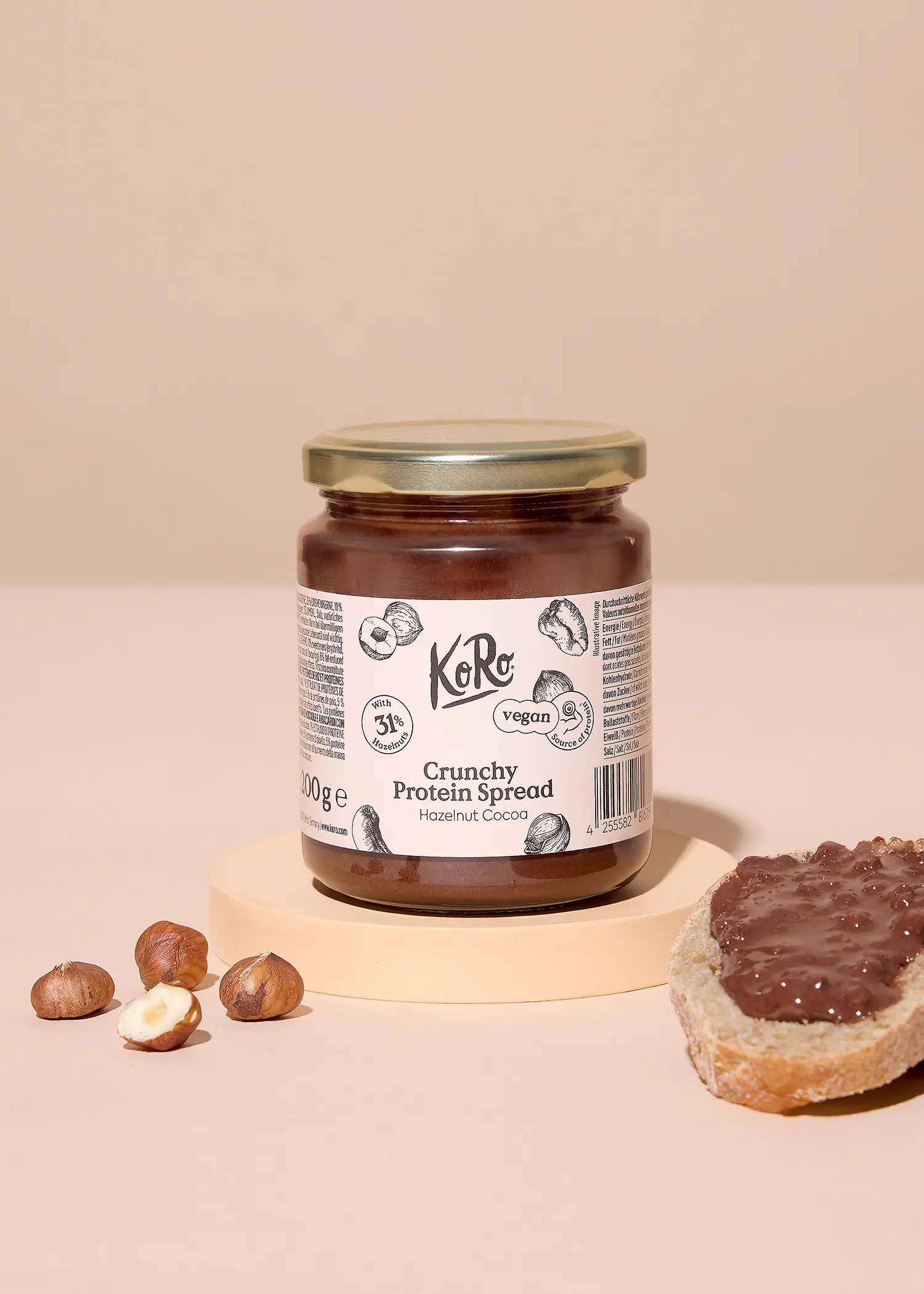 un pot de pâte à tartiner protéinée croquante ko ro noisette cacao, avec 31% de noisettes et végétalien, est posé sur un support clair à côté de noisettes entières et d'une tranche de pain tartinée.