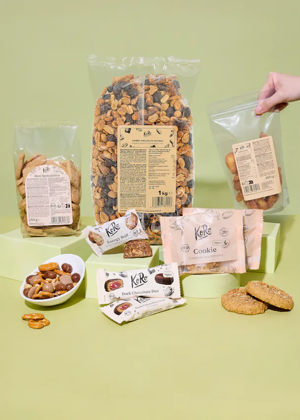 eine auswahl an koro-snacks, darunter eine große tüte süßer und salziger nussmischung, mini-spekulatius, apfelbäckchen, energiekugeln, dunkle schokoladenriegel und karamellisierte mandelcookies, präsentiert auf einem hellgrünen hintergrund.