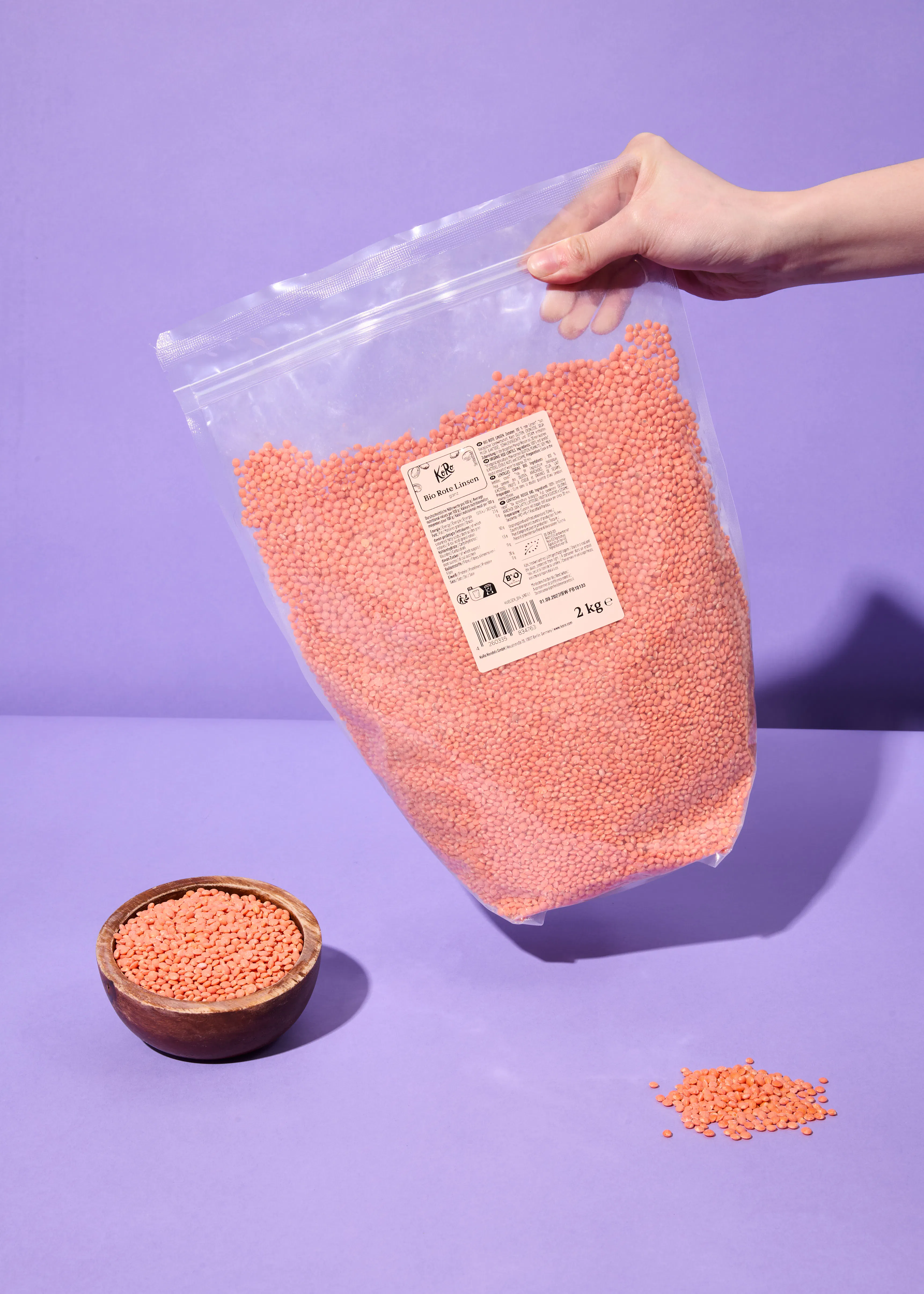 une main tient un grand sac transparent de 2 kg de lentilles corail bio, avec un petit bol en bois rempli de lentilles et une petite pile de lentilles éparpillées sur une surface violette.