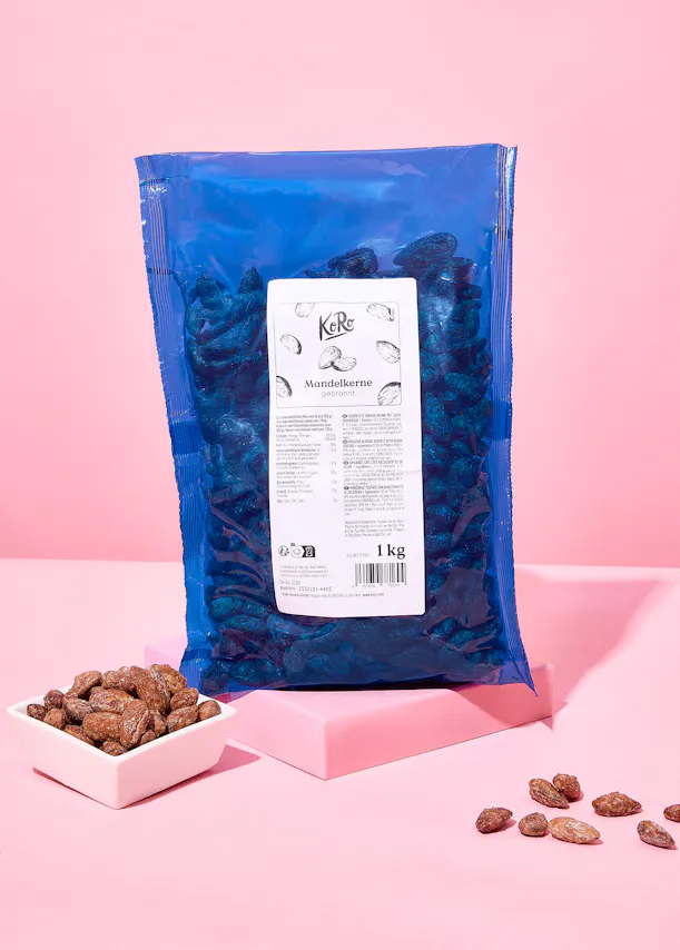 un grand sac bleu de 1 kg d'amandes grillées et sucrées de la marque koro, accompagné d'un petit bol rempli et de quelques amandes éparpillées sur une surface rose.
