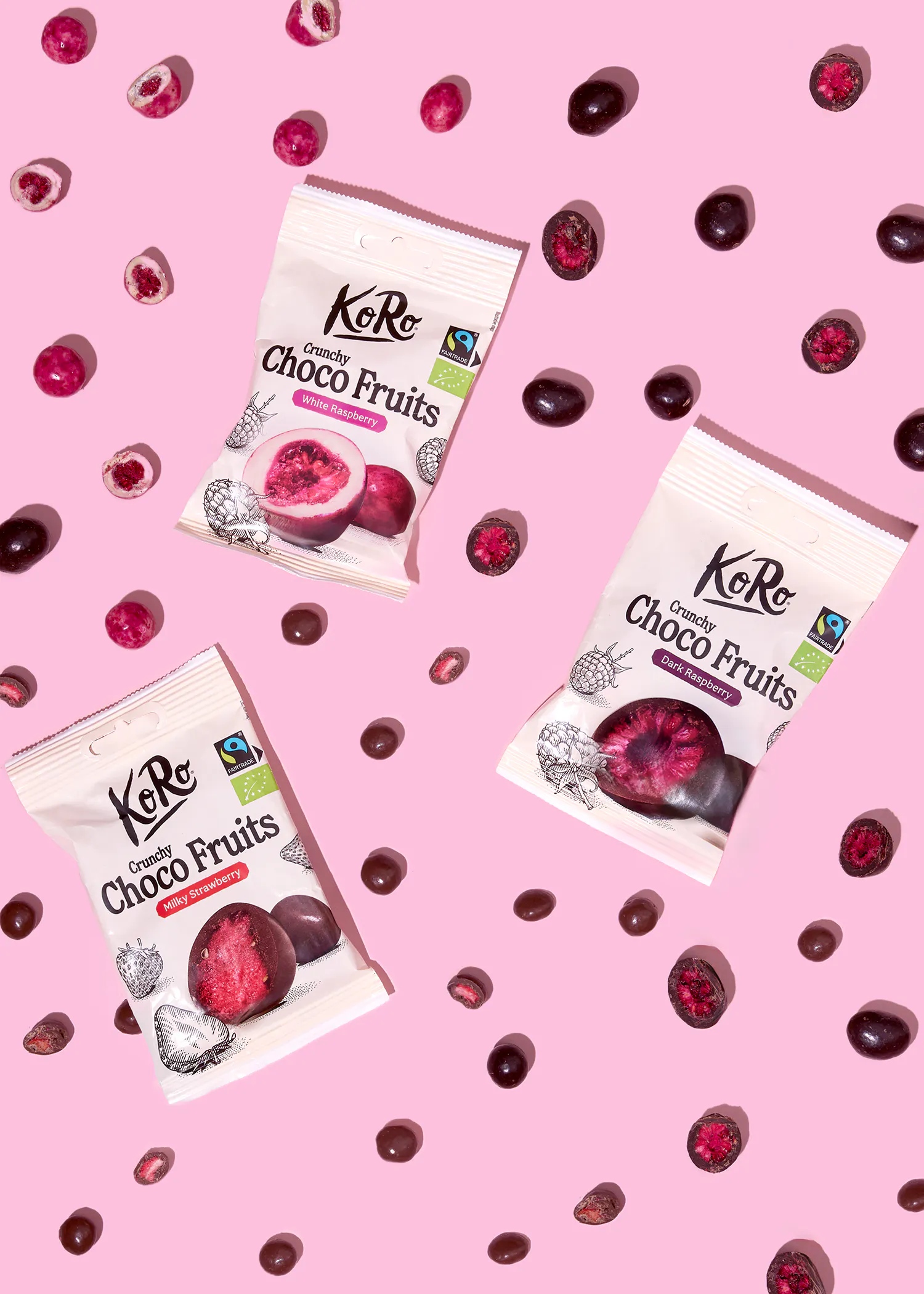 trois paquets de ko ro crunchy choco fruits (framboise blanche, fraise au lait et framboise noire) sont disposés sur un fond rose, entourés de fruits enrobés de chocolat.