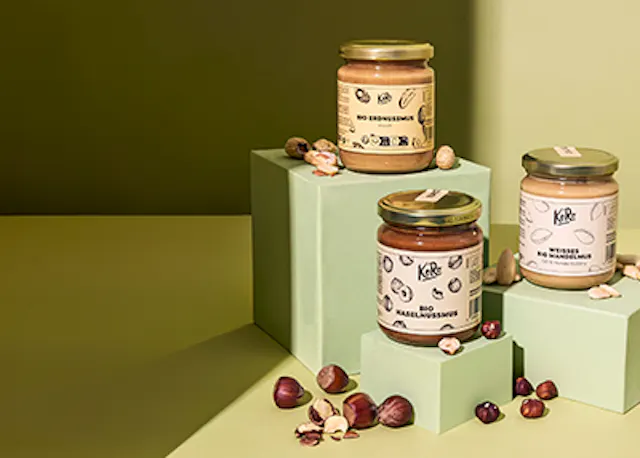 drei Gläser Haselnussmus stehen auf einem Tisch neben Haselnüssen