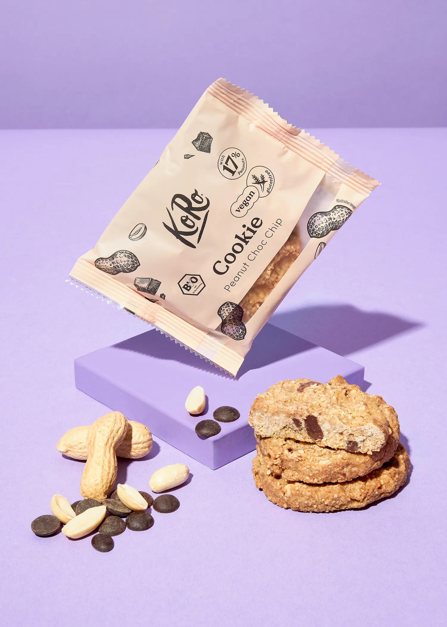 un paquet de biscuits koro aux pépites de chocolat et cacahuètes, végétalien et sans gluten, est posé sur un bloc violet à côté de biscuits, de cacahuètes et de pépites de chocolat sur un fond violet.