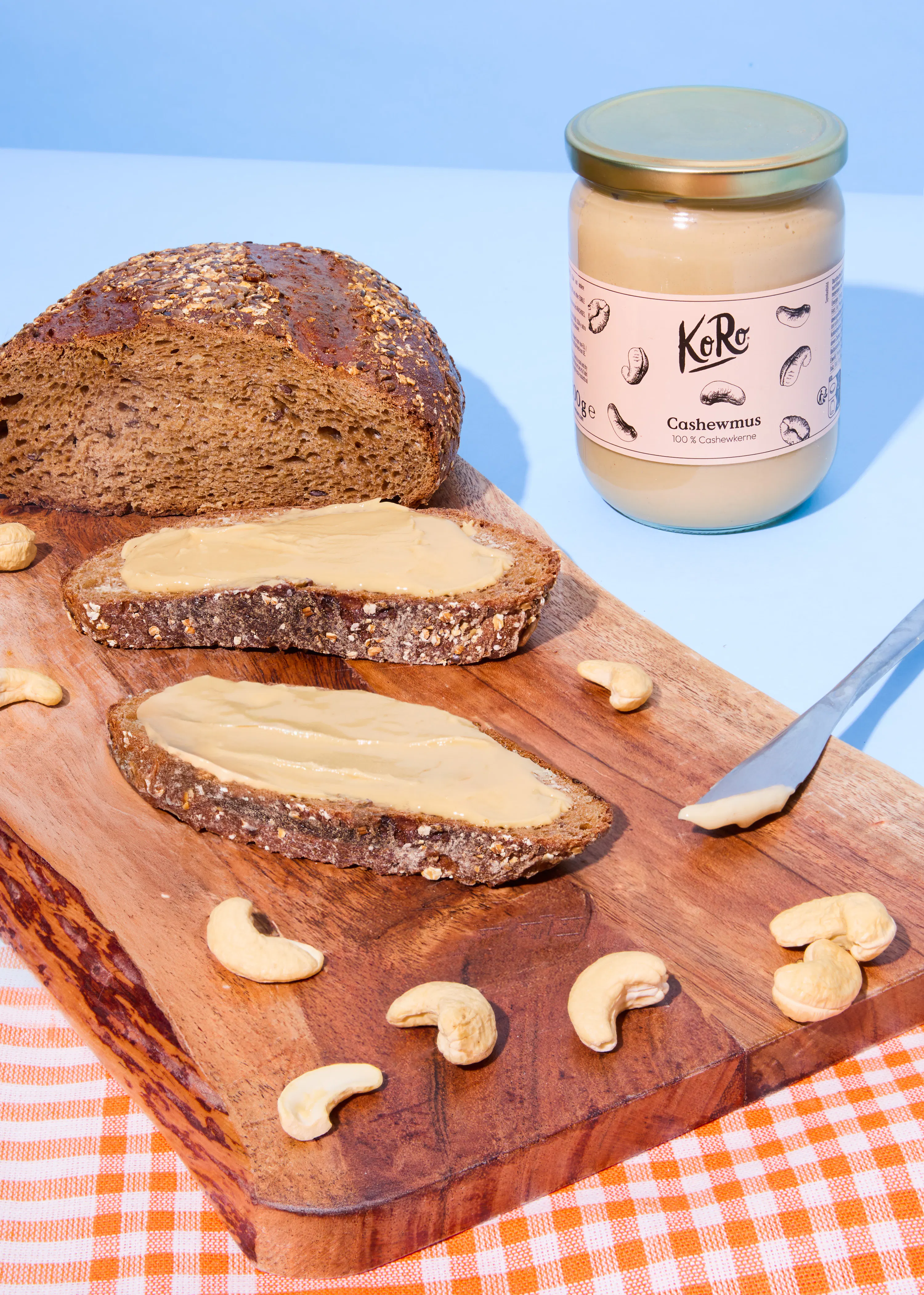 ein glas koro cashewnussmus steht neben einem laib vollkornbrot und zwei brotscheiben mit cashewnussmus, umgeben von ganzen cashewnüssen und einem messer auf einem holzbrett.