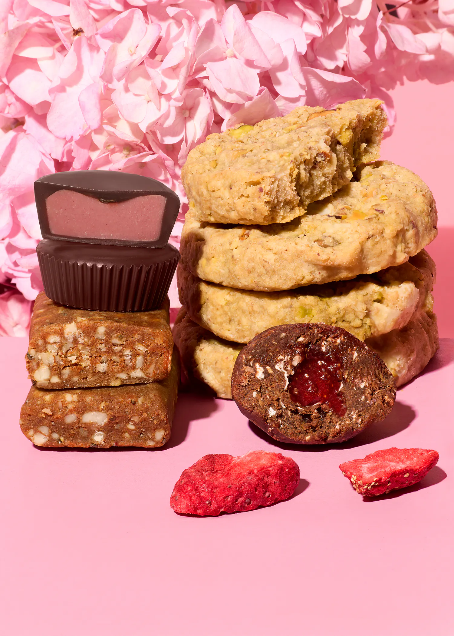 eine anordnung von süßigkeiten, darunter schokoladenpralinen mit pinker füllung, nussige kekse, fruchtriegel und eine gefüllte schokoladenkugel, zusammen mit getrockneten erdbeeren vor einem rosa blumenhintergrund.